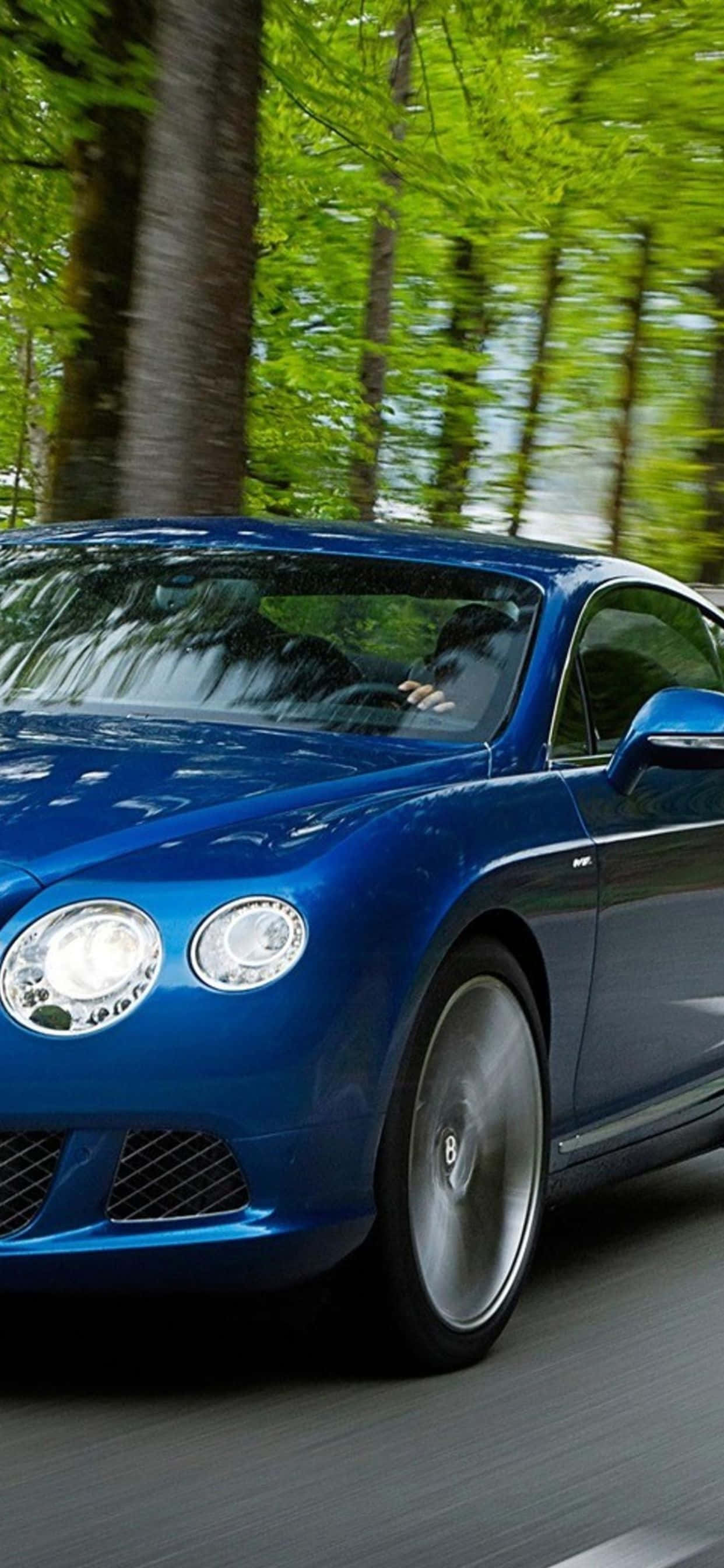 iPhone Xs Max Bentley 桌布 壁紙 — 一輛時尚的藍色賓利在陽光明媚的森林中疾馳，樹木和道路呈現動態模糊效果。
