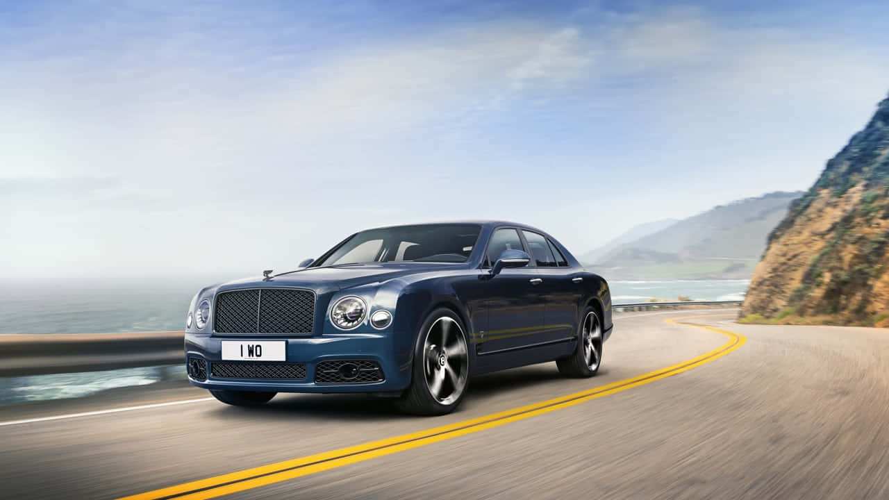 Bentley Mulsanne 桌布 壁紙 — 一輛時尚的藍色賓利轎車沿著海岸公路行駛，背景是山脈和海洋，天空晴朗。
