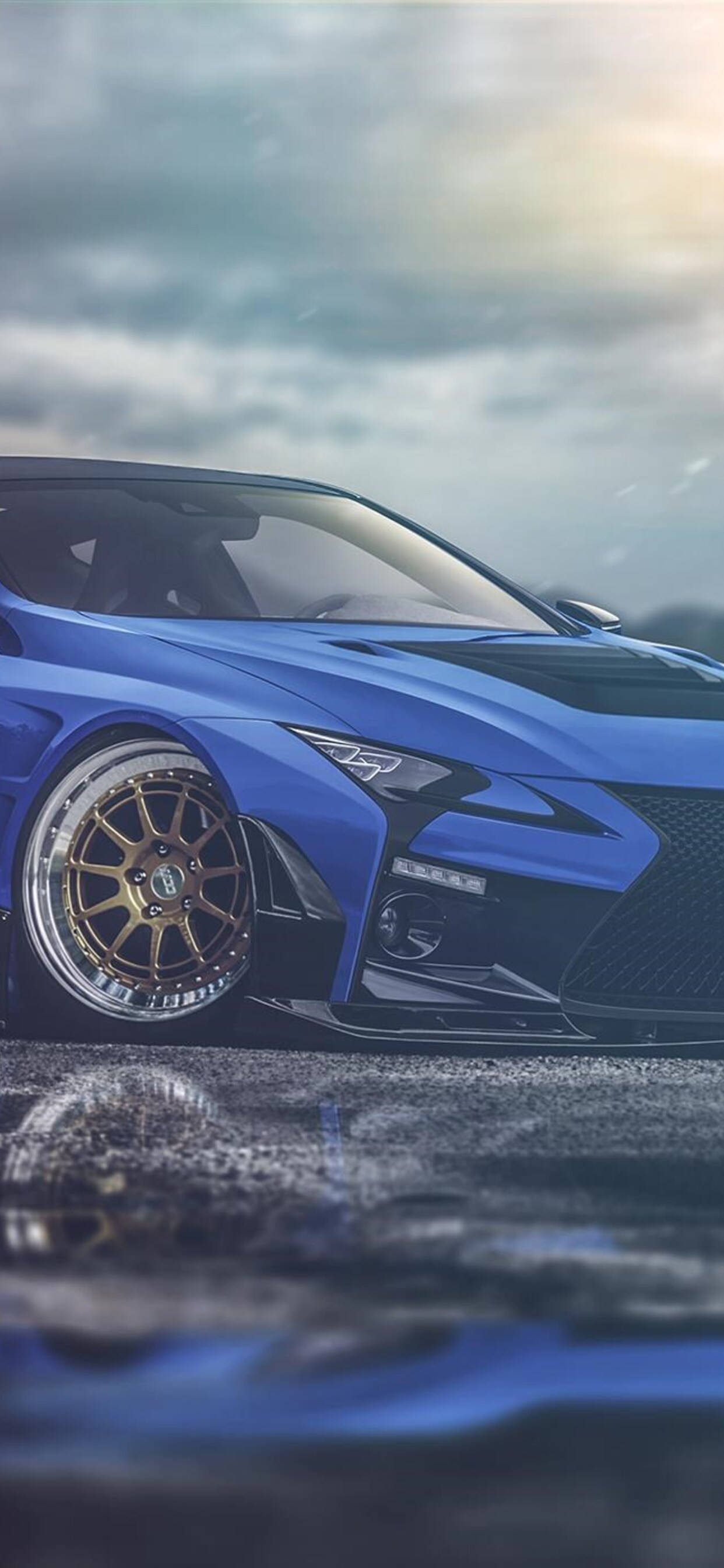lexus iPhone 桌布 壁紙 — 一輛時尚的藍色跑車配金色輪圈，低趴於潮濕路面上，天空多雲，帶有柔和的金色光線。