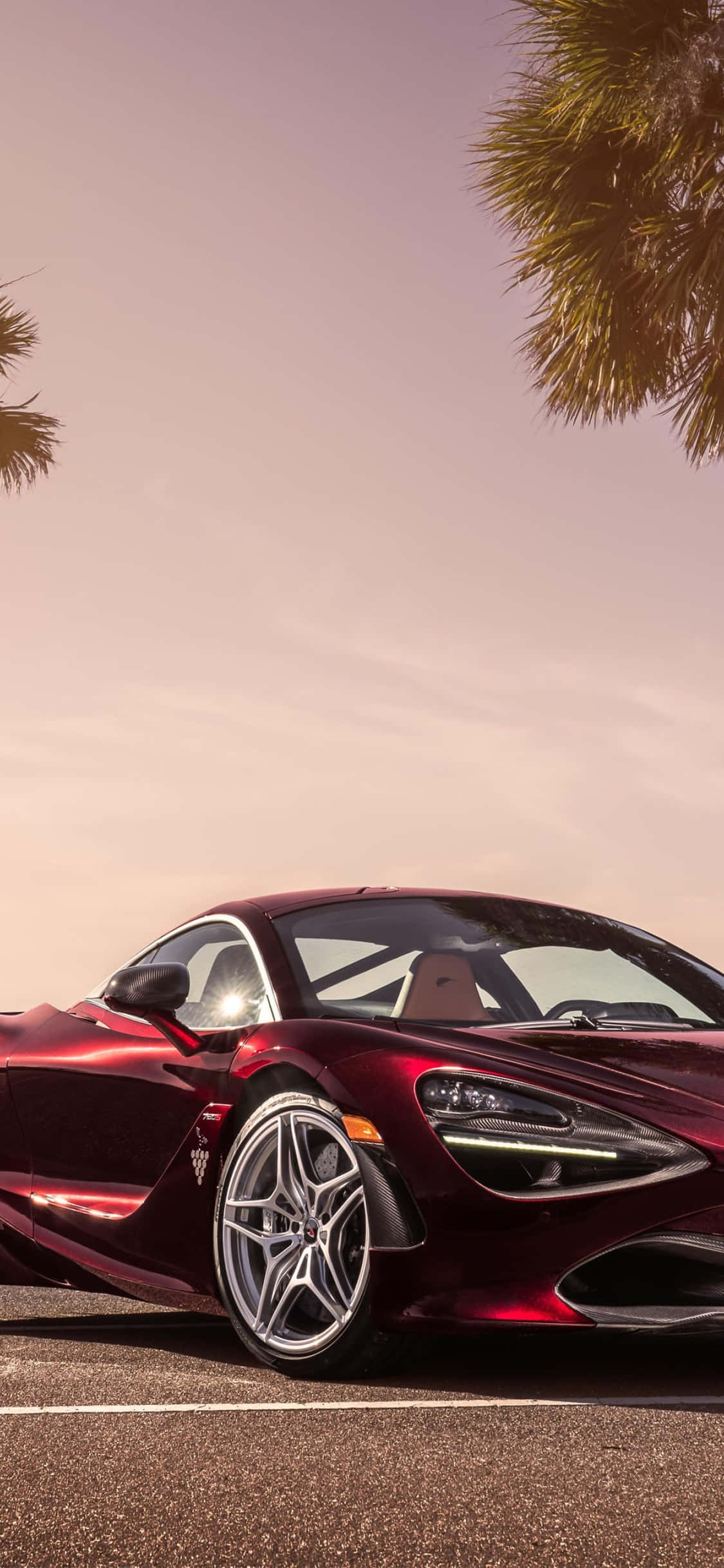 iPhone Xs Mclaren 720s 桌布 壁紙 — 一輛時尚的酒紅色跑車停在棕櫚樹下，夕陽西下，天空溫暖閃耀。