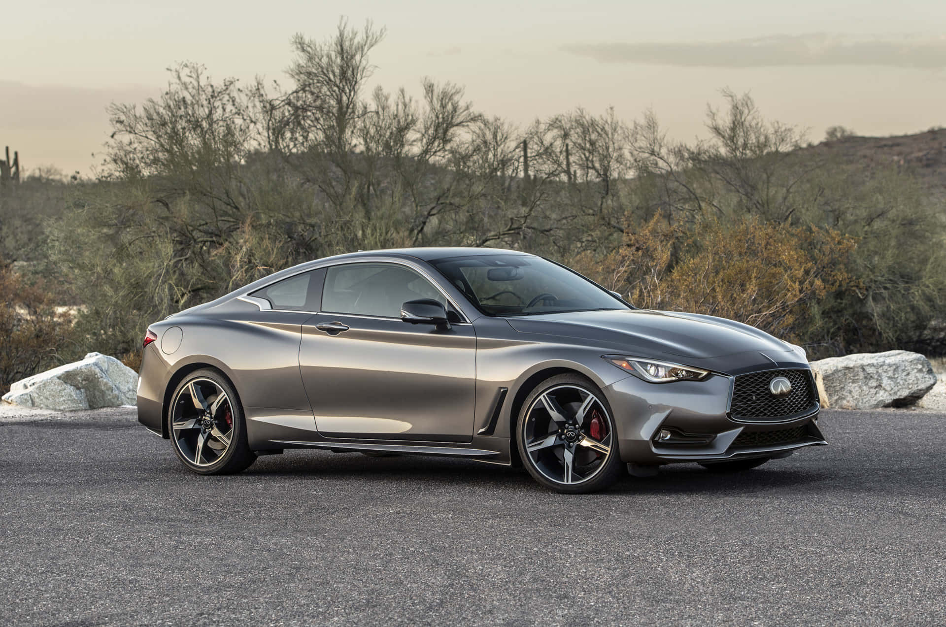 Infiniti Q60 桌布 壁紙 — 一輛時尚的金屬灰色運動轎跑車停在瀝青路上，背景是沙漠灌木和岩石，天空朦朧。