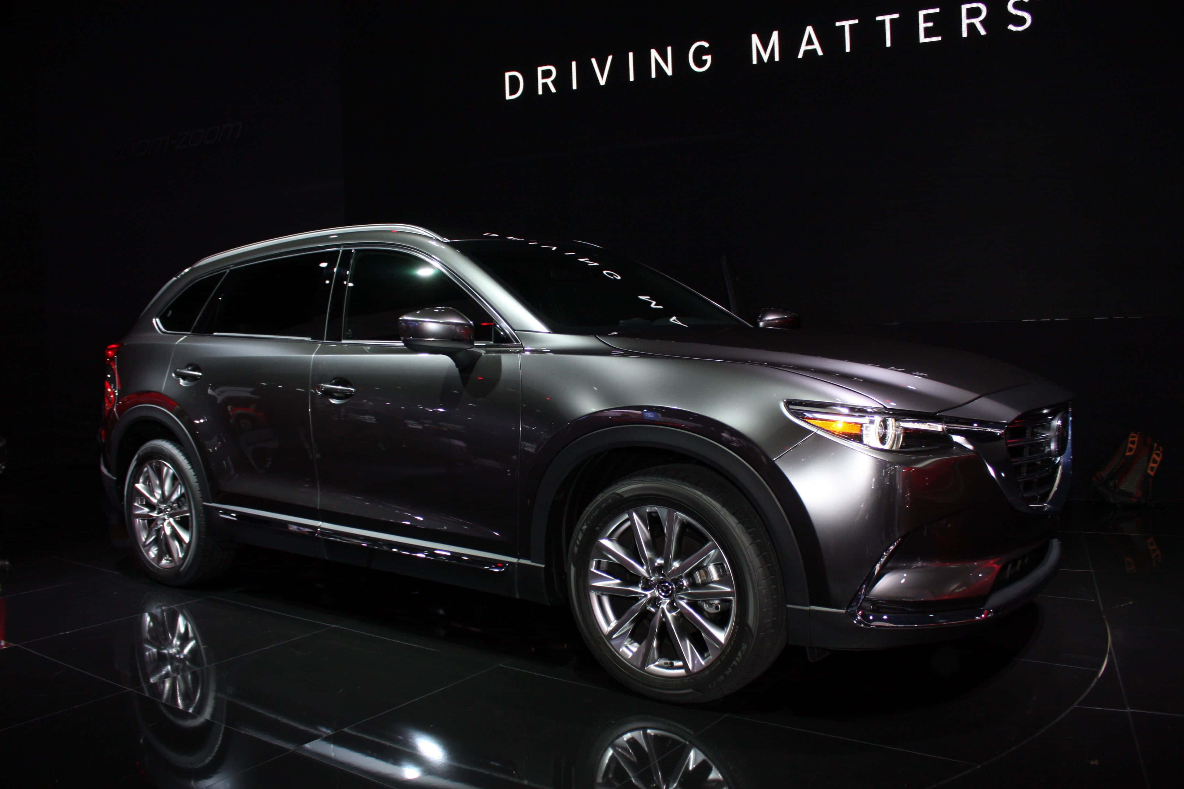 Mazda Cx-9 桌布 壁紙 — 一輛時尚的金屬灰色馬自達SUV在聚光燈下於反射黑色舞台上閃耀，上方有'DRIVING MATTERS'標牌。