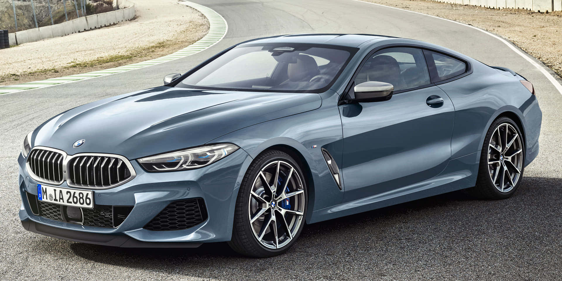 Bmw 8 Series 桌布 壁紙 — 一輛時尚的金屬藍色BMW 8系列轎跑車在明亮的日光下，沿著一條由礫石和綠樹環繞的彎曲瀝青道路滑行。