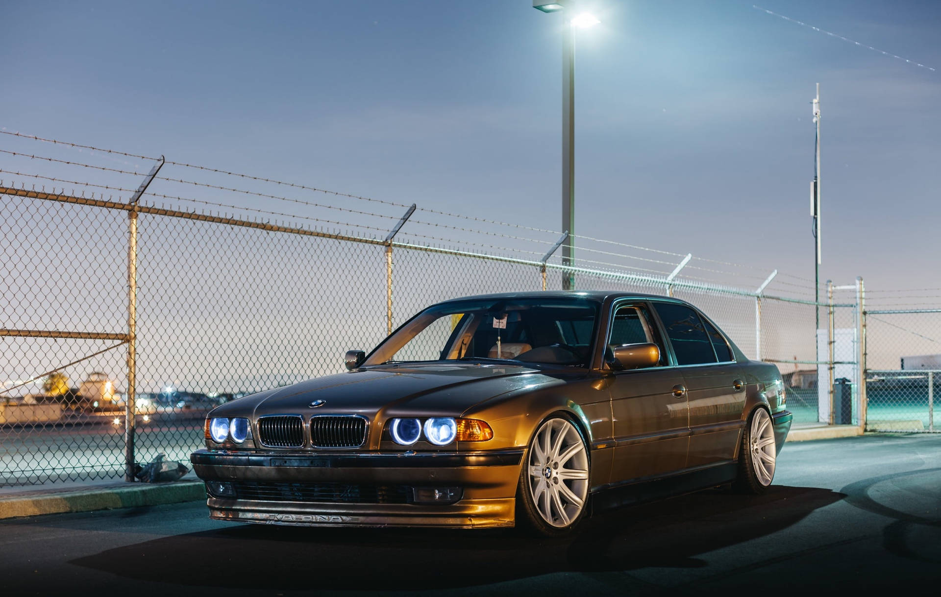 BMW 動圖 桌布 壁紙 — 一輛時尚的金色BMW轎車，車頭燈閃爍，在黃昏時停泊在明亮的街燈下的鐵絲網圍欄後面。