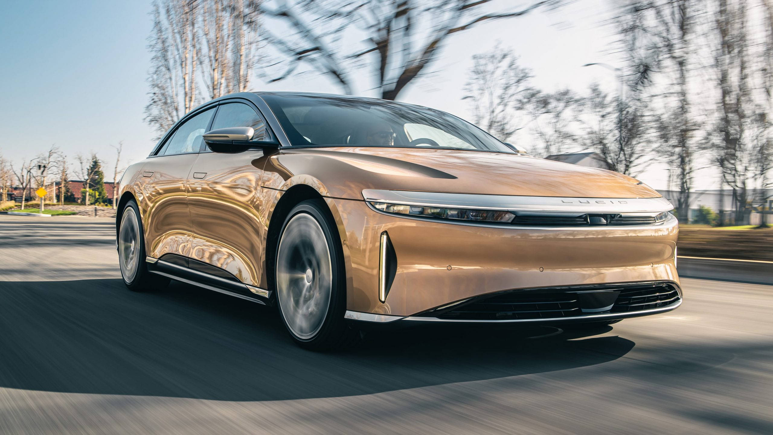 cars 桌布 壁紙 — 一輛時尚的金色電動車在郊區道路上疾馳，背景是模糊的樹木和晴朗的藍天。