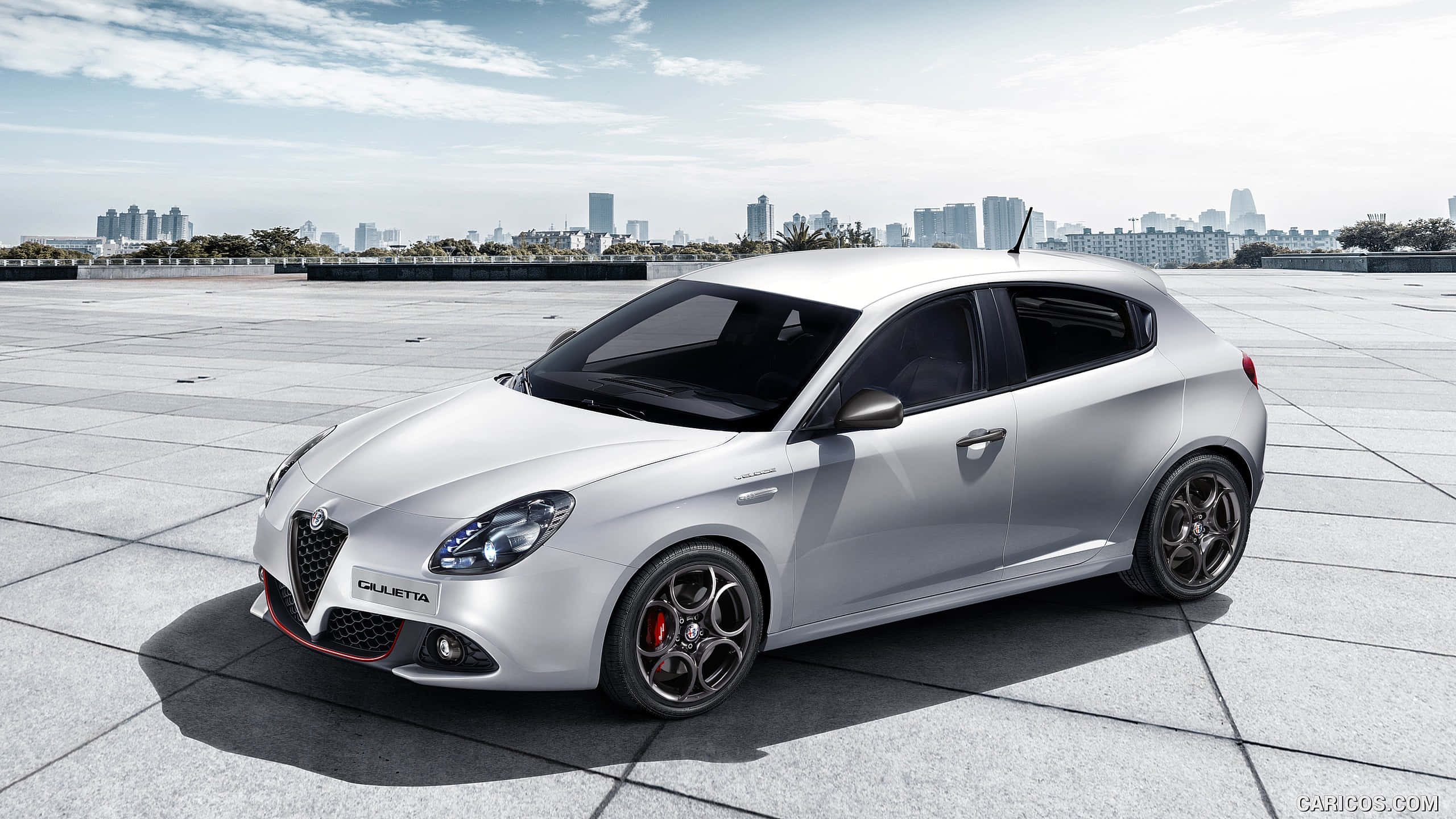 Alfa Romeo Giulietta 桌布 壁紙 — 一輛時尚的銀色 Alfa Romeo Giulietta 跑車停放在瓷磚屋頂上，背景是晴朗天空下的城市天際線。