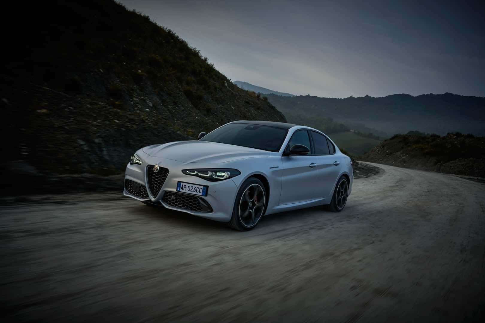愛快羅密歐 桌布 壁紙 — 一輛時尚的銀色 Alfa Romeo 轎車在蜿蜒的山路上疾馳，天空籠罩著暮色氛圍。