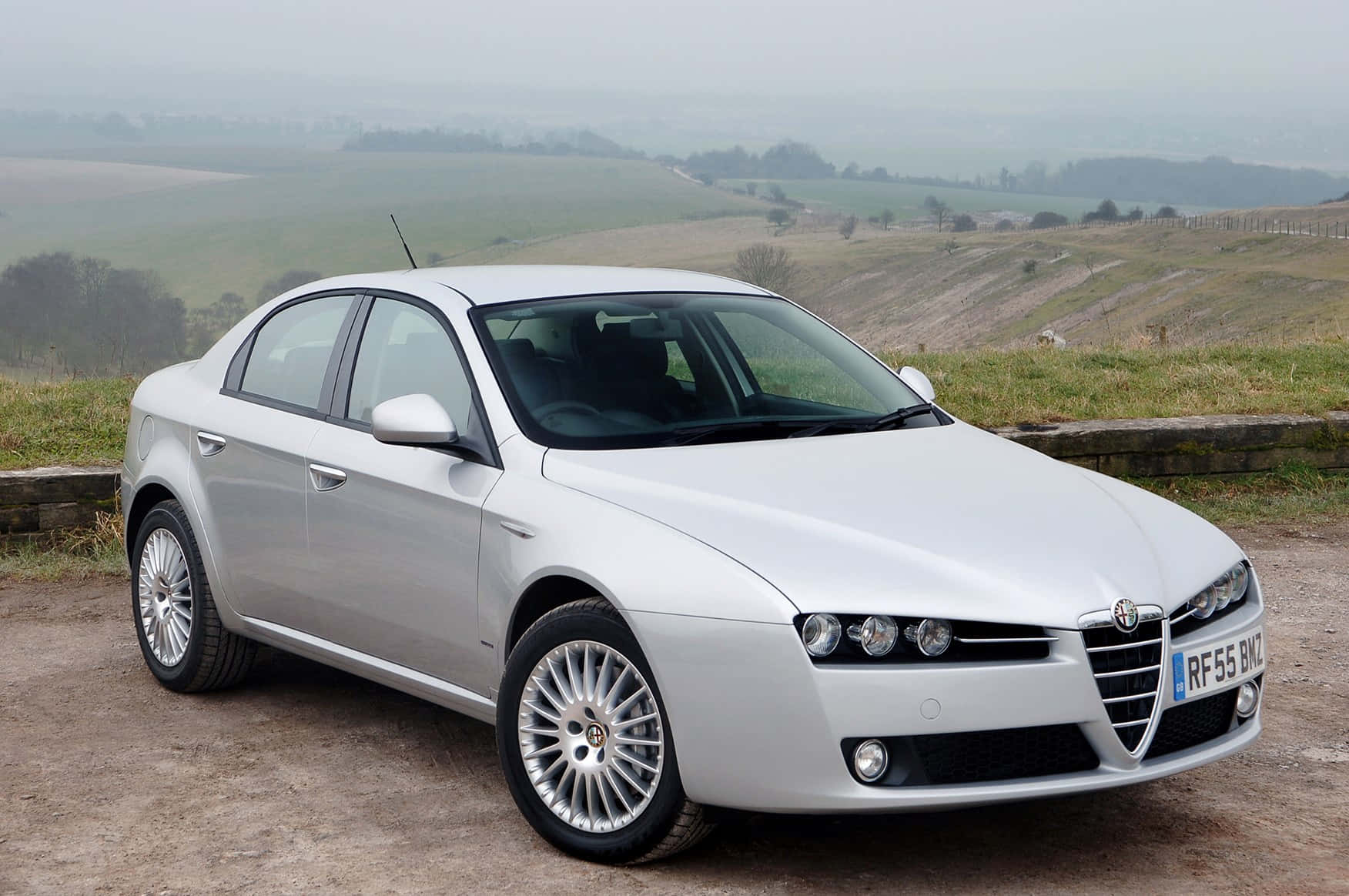 Alfa Romeo 159 桌布 壁紙 — 一輛時尚的銀色 Alfa Romeo 轎車停泊在碎石路上，俯瞰著煙霧繚繞的連綿綠色山丘。
