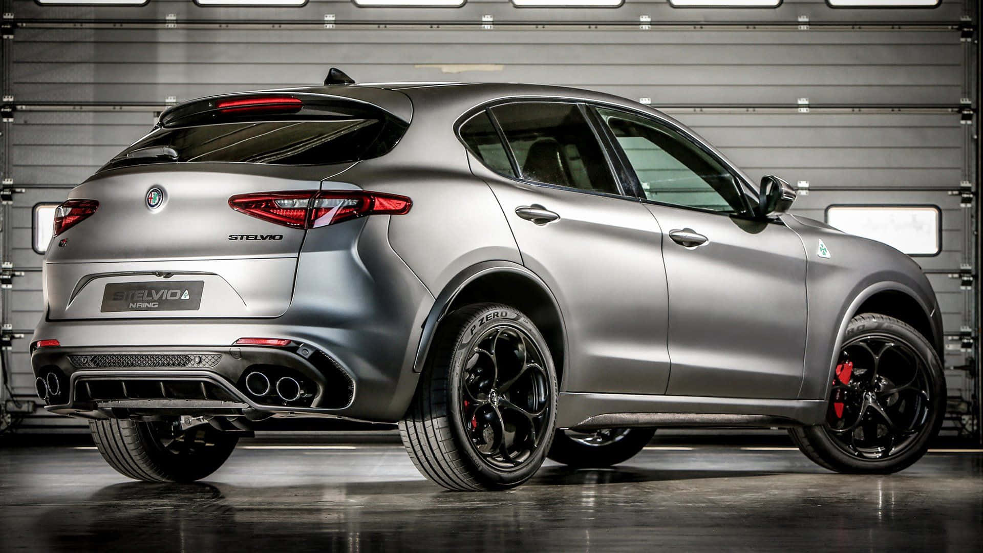 Alfa Romeo Stelvio 桌布 壁紙 — 一輛時尚的銀色 Alfa Romeo Stelvio Quadrifoglio SUV，配備黑色輪圈和紅色制動卡鉗，在帶有反射地板的現代車庫中展示。