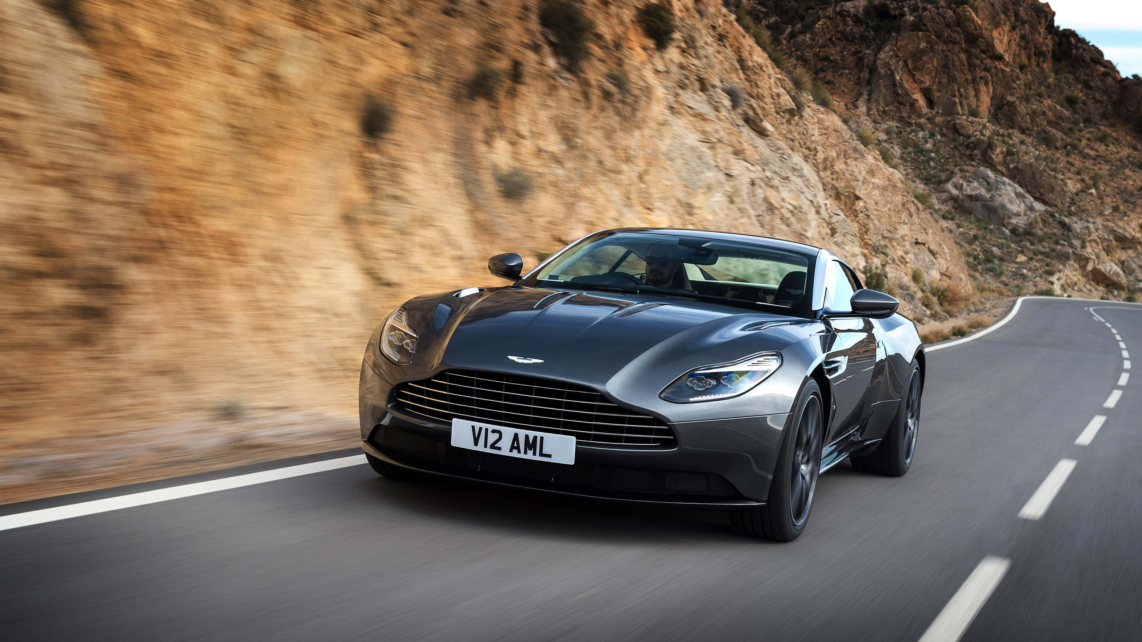 奧斯頓馬丁 桌布 壁紙 — 一輛時尚的銀色 Aston Martin V12 AMI 沿著蜿蜒的山路疾馳，模糊的岩石懸崖呼嘯而過。