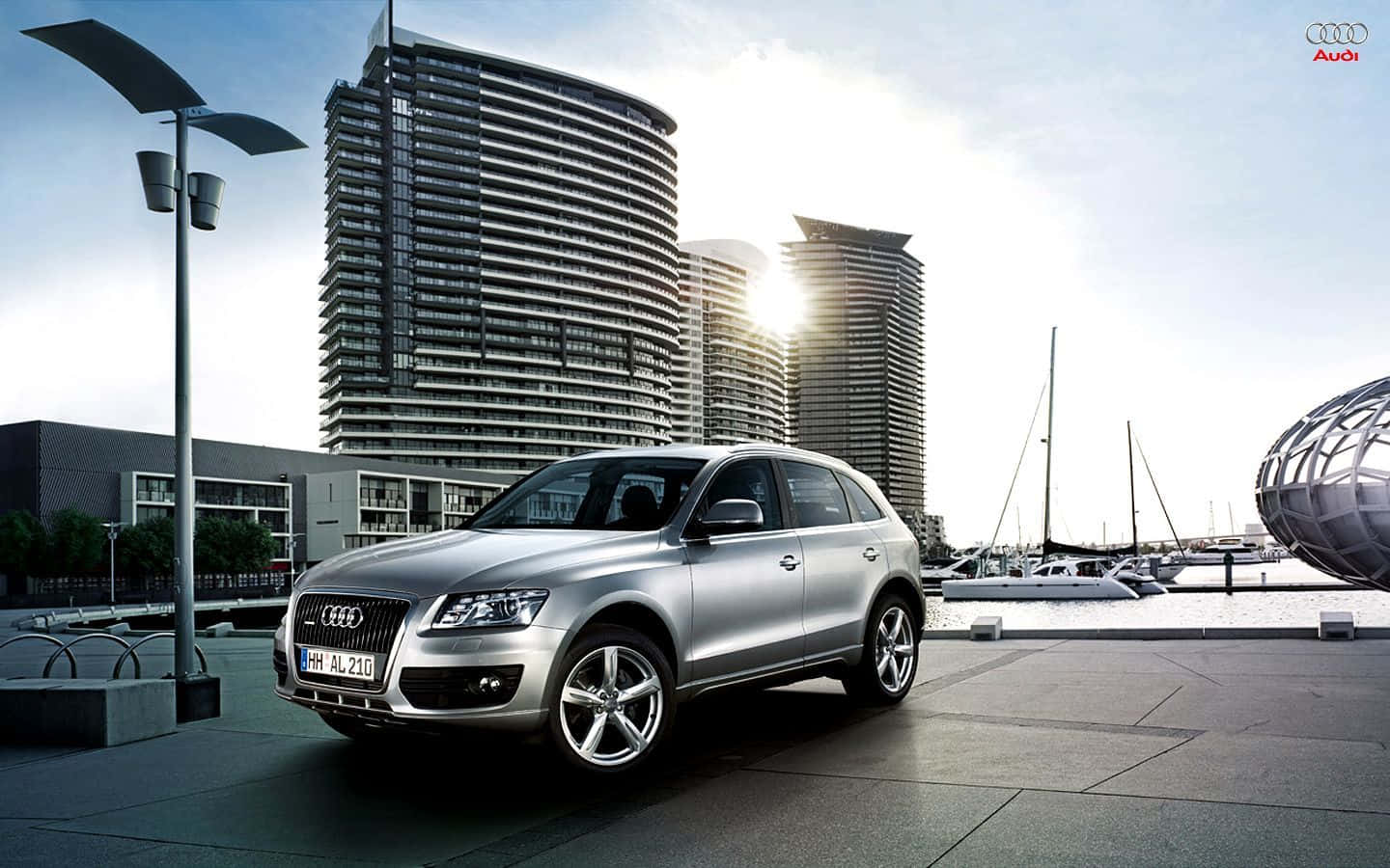 Audi Sq5 桌布 壁紙 — 一輛時尚的銀色 Audi Q7 SUV 停泊在現代海濱廣場上，背景有彎曲的摩天大樓和帆船。