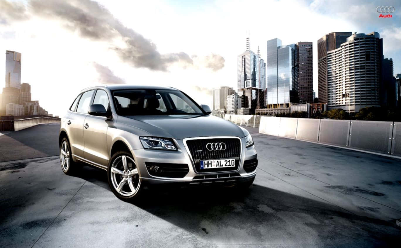Audi Sq5 桌布 壁紙 — 一輛時尚的銀色 Audi Q7 SUV 停泊在混凝土天橋上，背景是現代城市天際線和戲劇性的雲層。