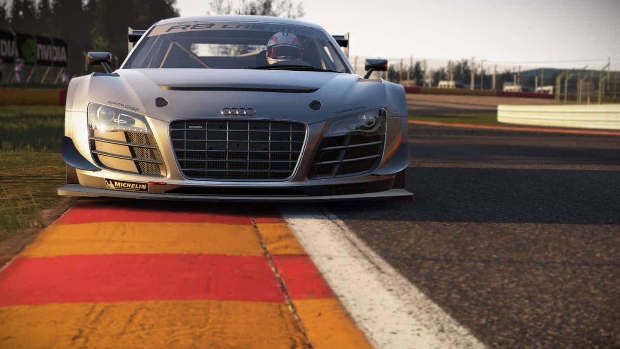 電玩遊戲 桌布 壁紙 — 一輛時尚的銀色 Audi R8 LMS 賽車停在賽道邊緣，賽道上有紅色和黃色的路緣石，在溫暖的日光下。