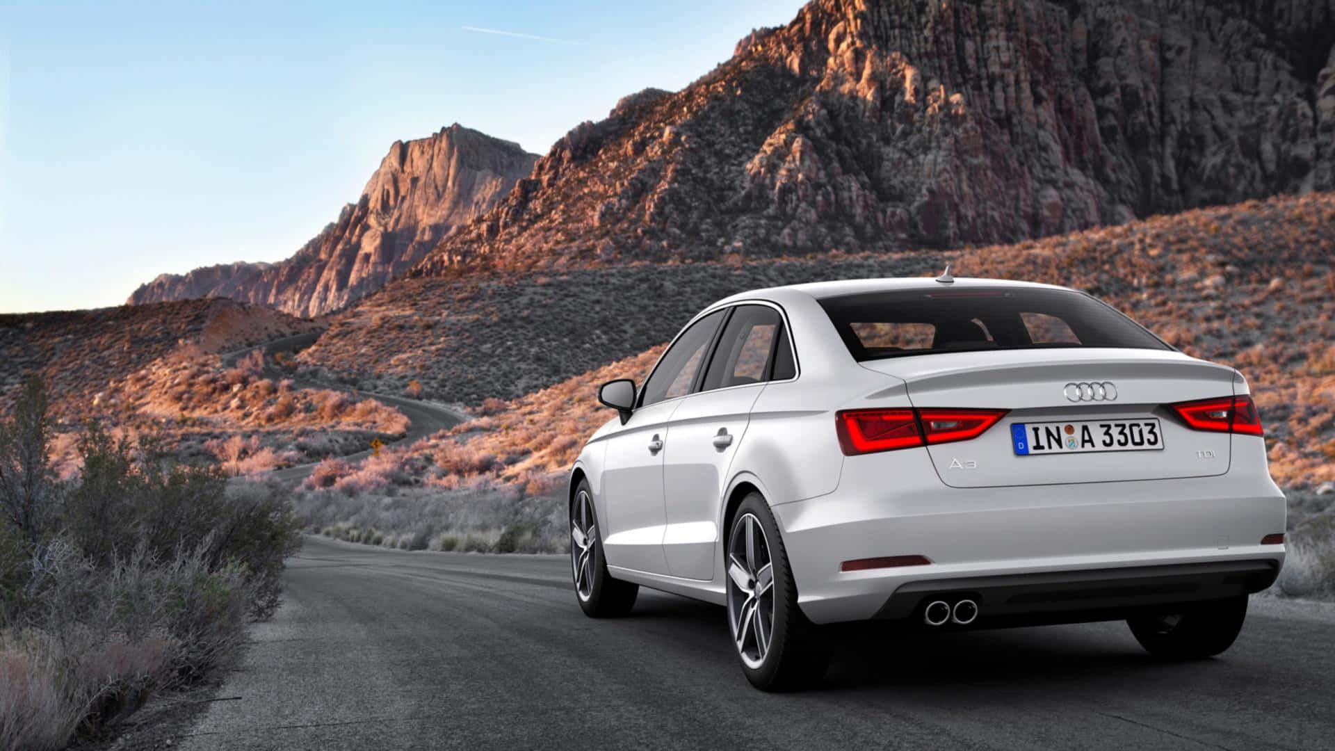 Audi A3 桌布 壁紙 — 一輛時尚的銀色 Audi 房車沿著蜿蜒的沙漠道路行駛，崎嶇的山脈沐浴在金色的日落光芒中。