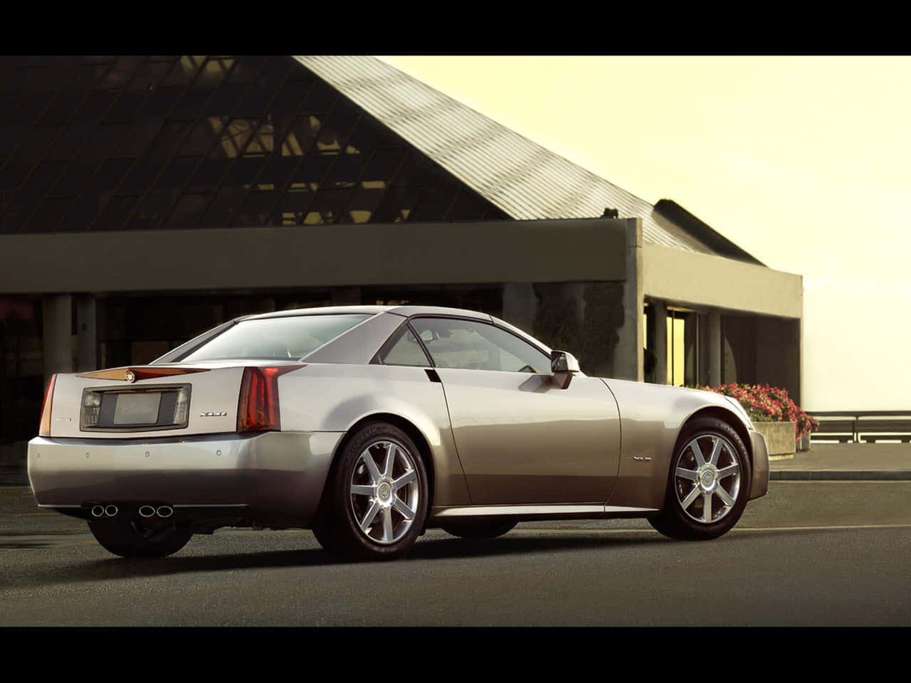 Cadillac Xlr 桌布 壁紙 — 一輛時尚的銀色 Cadillac XLR 雙門跑車停在柏油路面上，旁邊是一座帶有斜屋頂和花槽的現代建築。