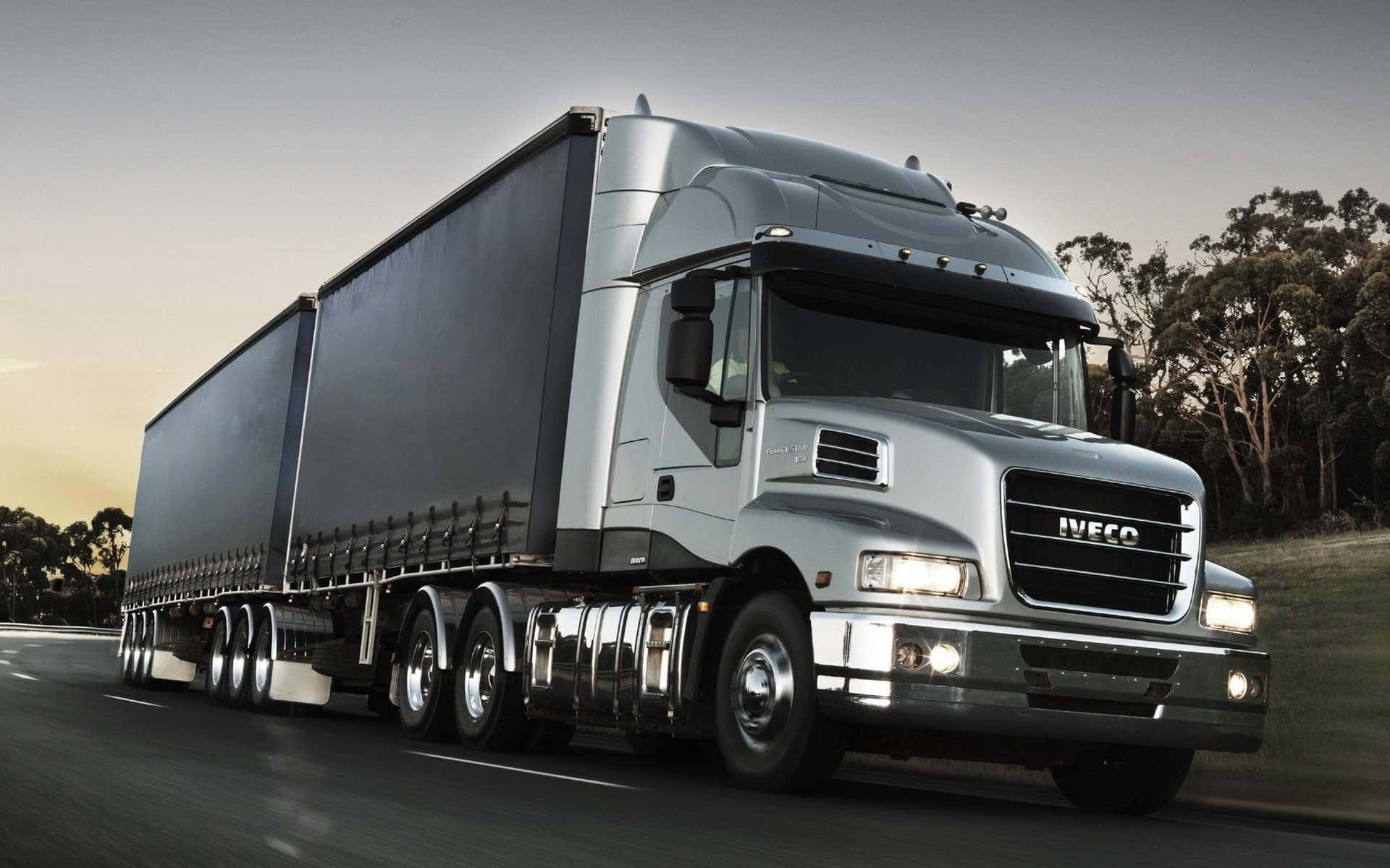 Big Truck 桌布 壁紙 — 一輛時尚的銀色 Iveco 半聯結卡車拖著黑色拖車，在日落時分行駛在高速公路上，背景有樹木。