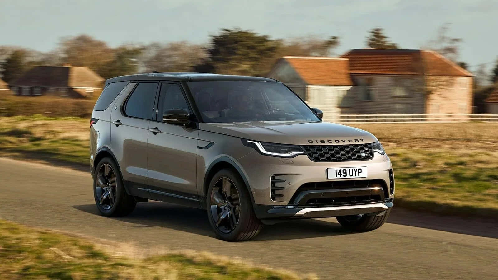Land Rover Discovery 桌布 壁紙 — 一輛時尚的銀色 Land Rover Discovery 沿著鄉間道路行駛，經過質樸的石屋，天空晴朗。