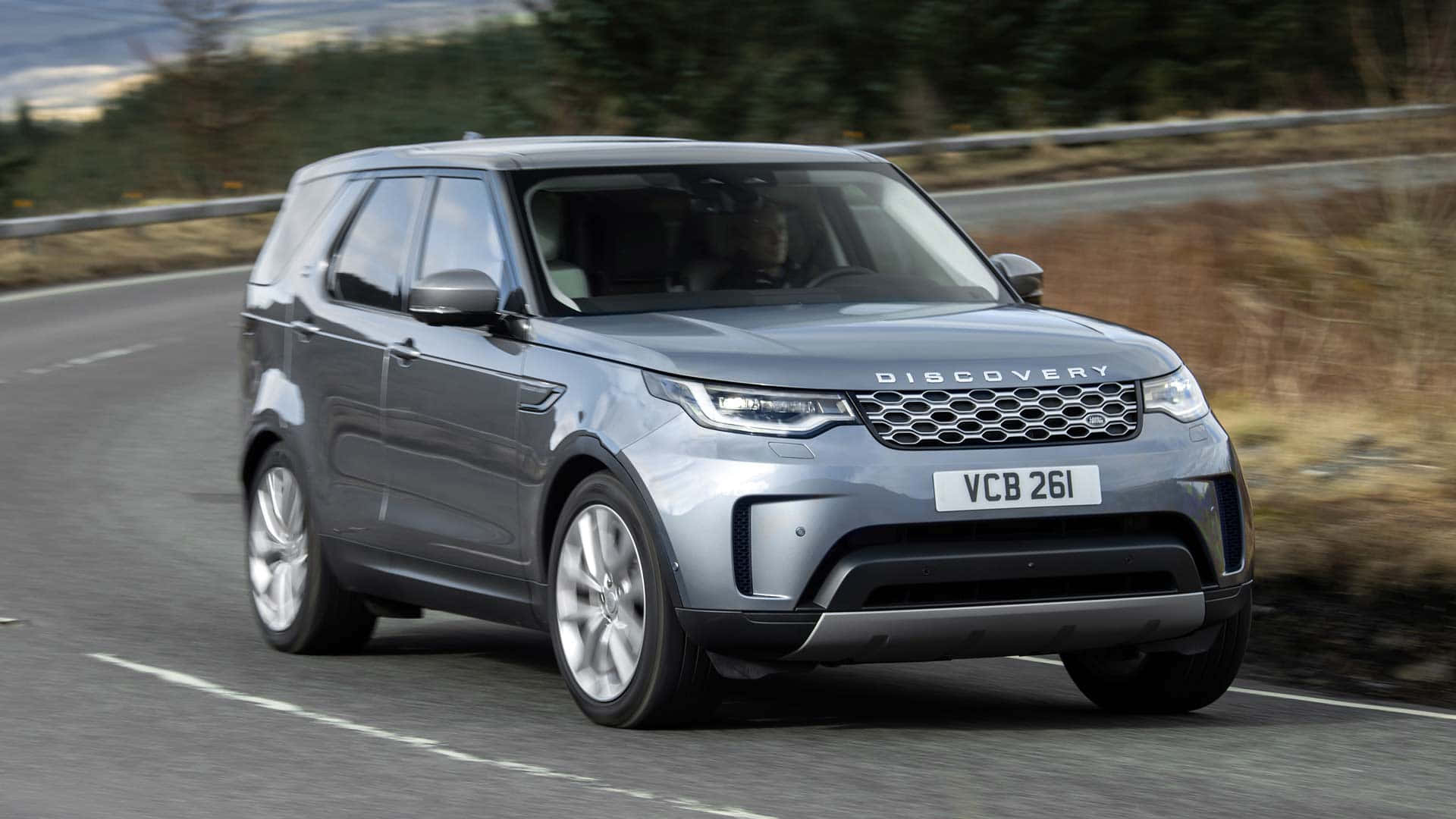 Land Rover Discovery 桌布 壁紙 — 一輛時尚的銀色 Land Rover Discovery 沿著蜿蜒的道路行駛，穿過模糊的自然景觀。