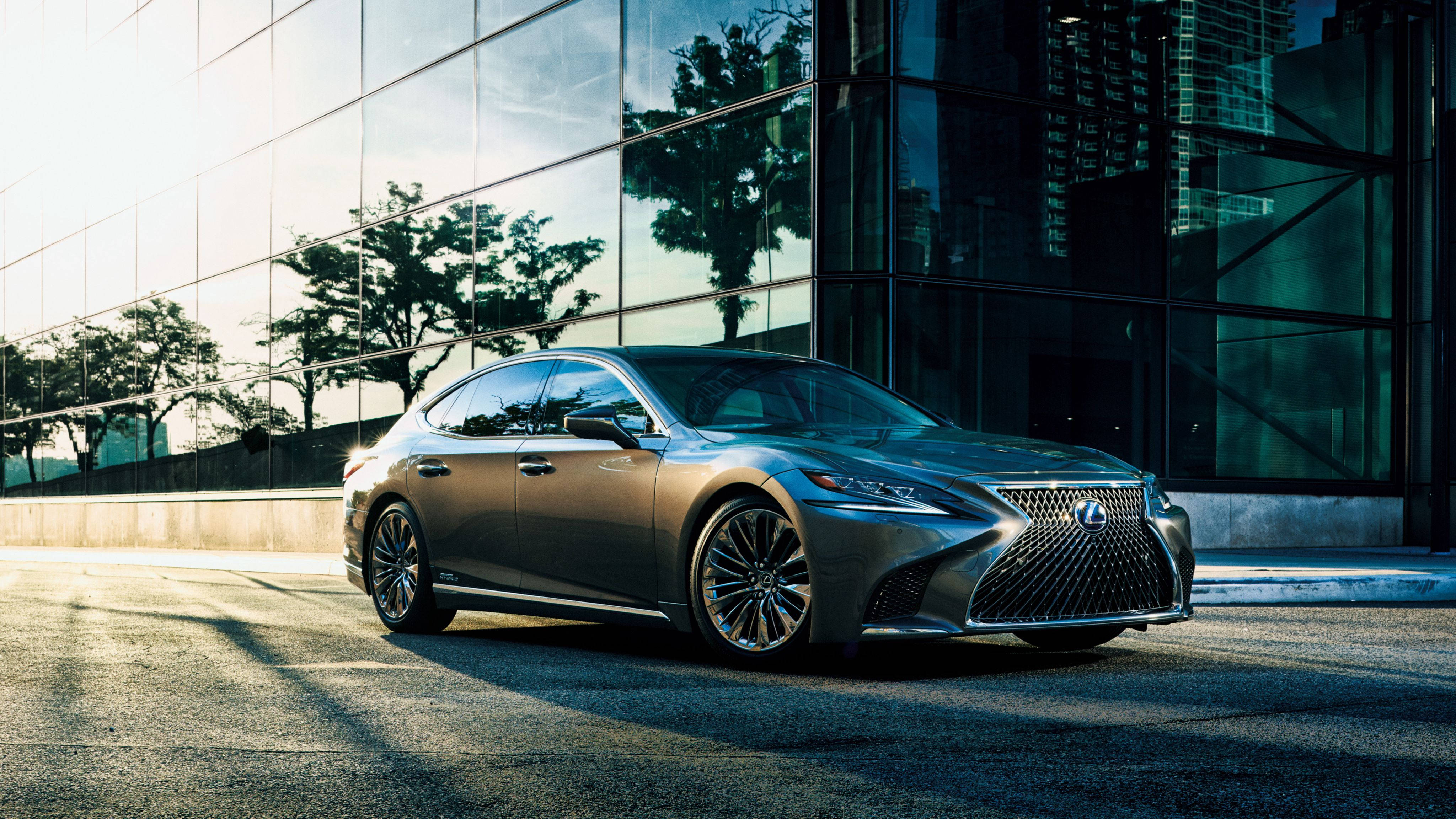 Lexus 桌布 壁紙 — 一輛時尚的銀色 Lexus 房車停泊在陽光明媚的城市街道上，旁邊是一座反射樹木的玻璃建築。