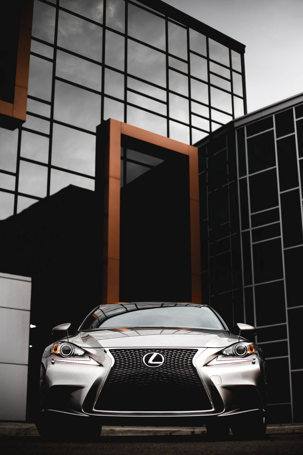 Lexus 標誌 桌布 壁紙 — 一輛時尚的銀色 Lexus 跑車停放在現代玻璃和鋼鐵建築前，天空多雲。