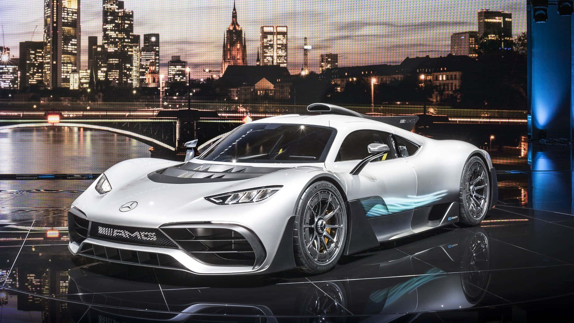 Mercedes 桌布 壁紙 — 一輛時尚的銀色 Mercedes-AMG 跑車在舞台燈光下閃閃發光，背景是黃昏時分充滿活力的城市天際線。