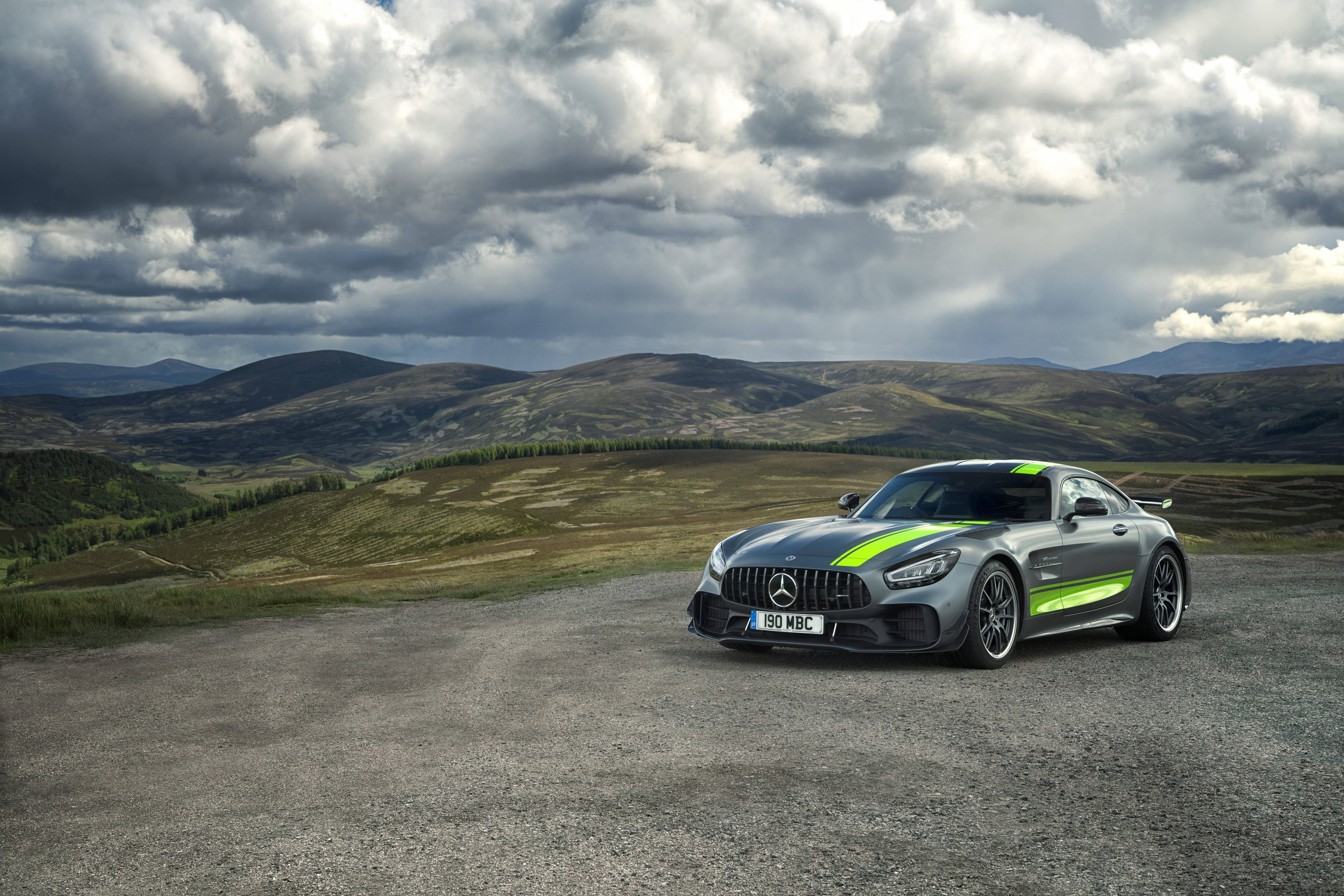 Gtr 桌布 壁紙 — 一輛時尚的銀色 Mercedes-AMG GT 帶有螢光綠色條紋，停在碎石路上，背景是戲劇性的多雲天空和起伏的山丘。