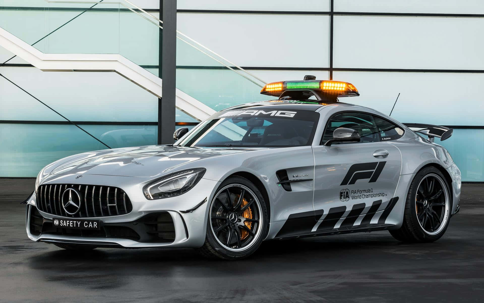 Safety Car 桌布 壁紙 — 一輛時尚的銀色 Mercedes-AMG GT 安全車，帶有閃爍的燈光，停在一棟現代玻璃建築前。