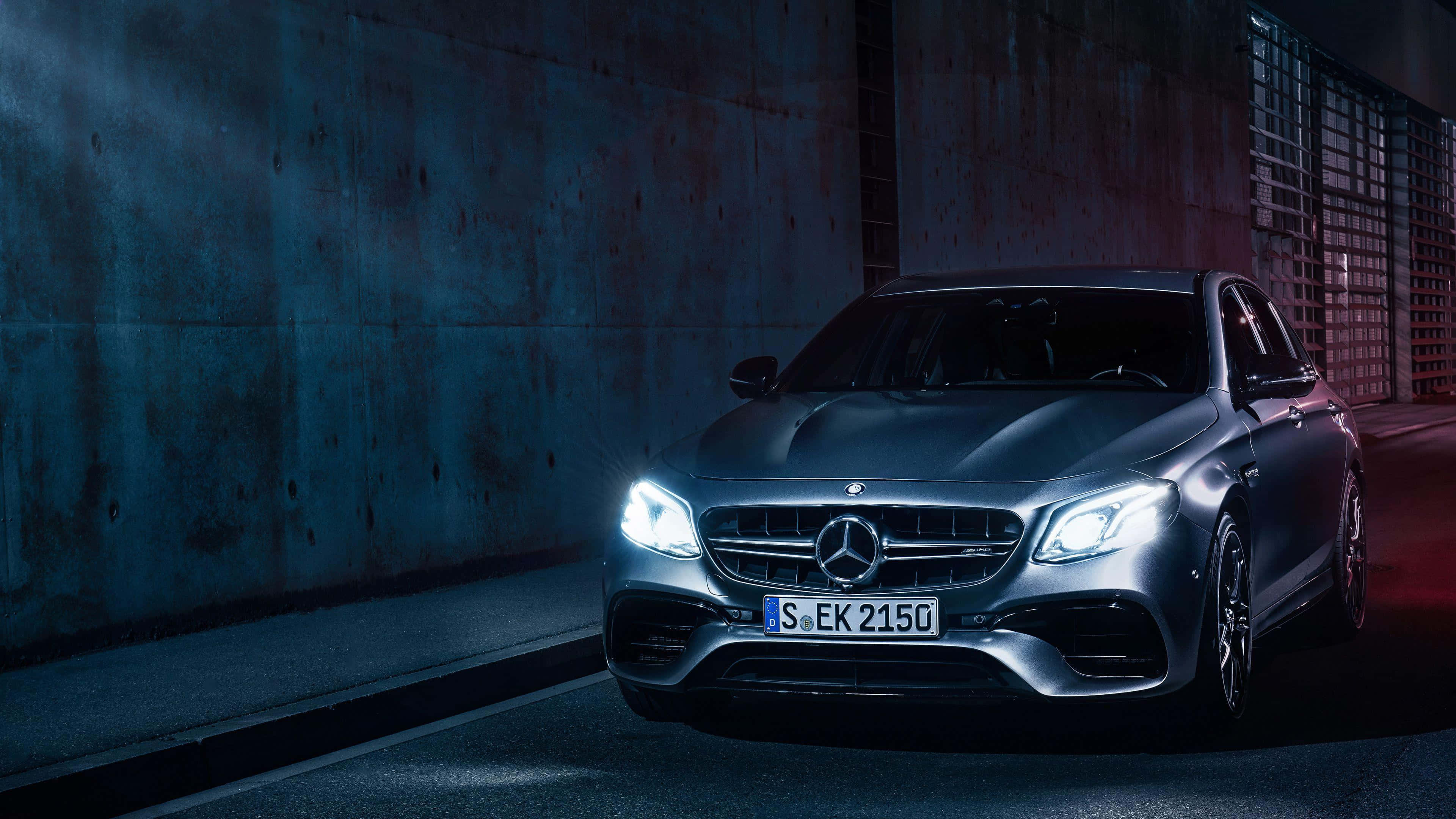 Mercedes 桌布 壁紙 — 一輛時尚的銀色 Mercedes-AMG E63 S 轎車，配備發光頭燈，停在光線昏暗的城市隧道中，帶有藍色和紅色的燈光點綴。