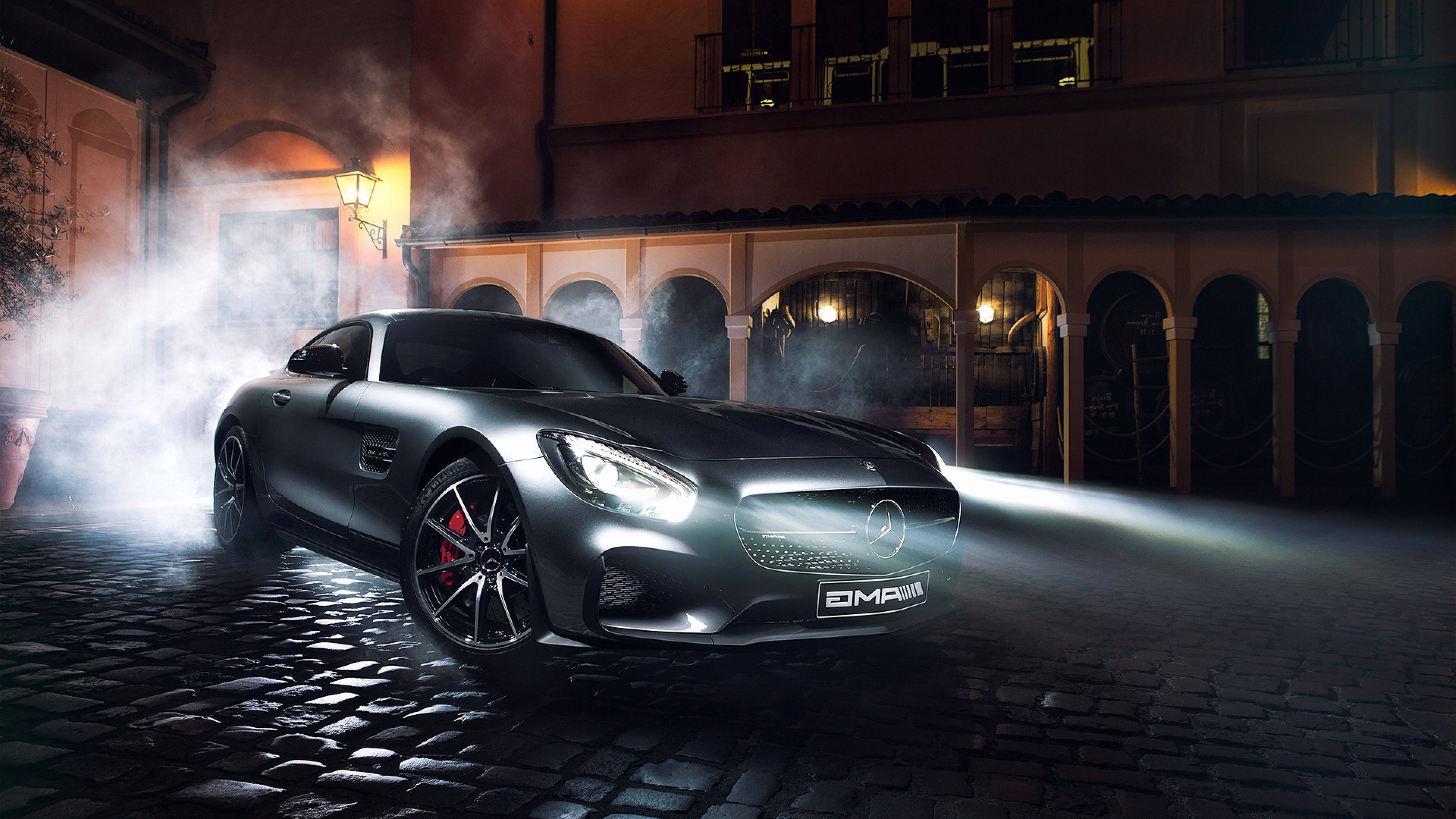 Mercedes 桌布 壁紙 — 一輛時尚的銀色 Mercedes-AMG GT 跑車，車頭燈閃爍，在夜間穿過霧氣籠罩的鵝卵石街道。