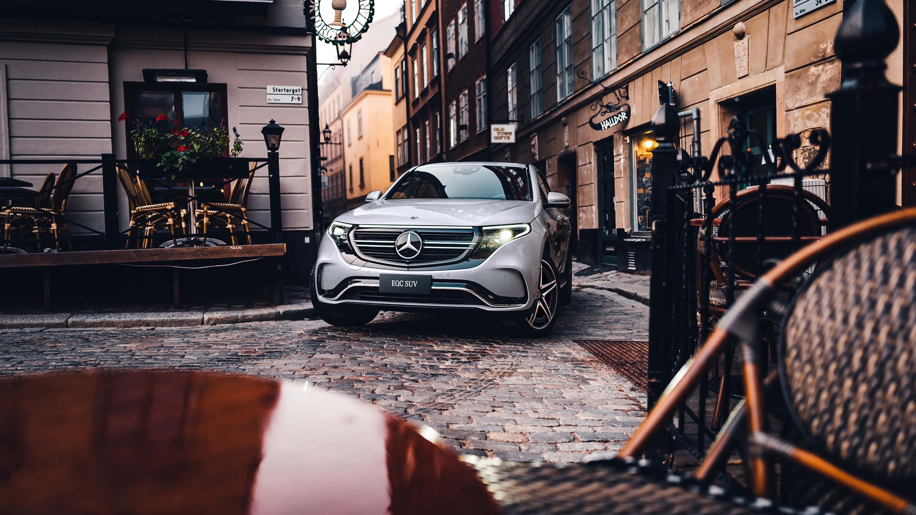 Mercedes 桌布 壁紙 — 一輛時尚的銀色 Mercedes-Benz EQC SUV 停泊在歐洲鵝卵石街道上，正值黃金時段，旁邊有咖啡桌和歷史建築。
