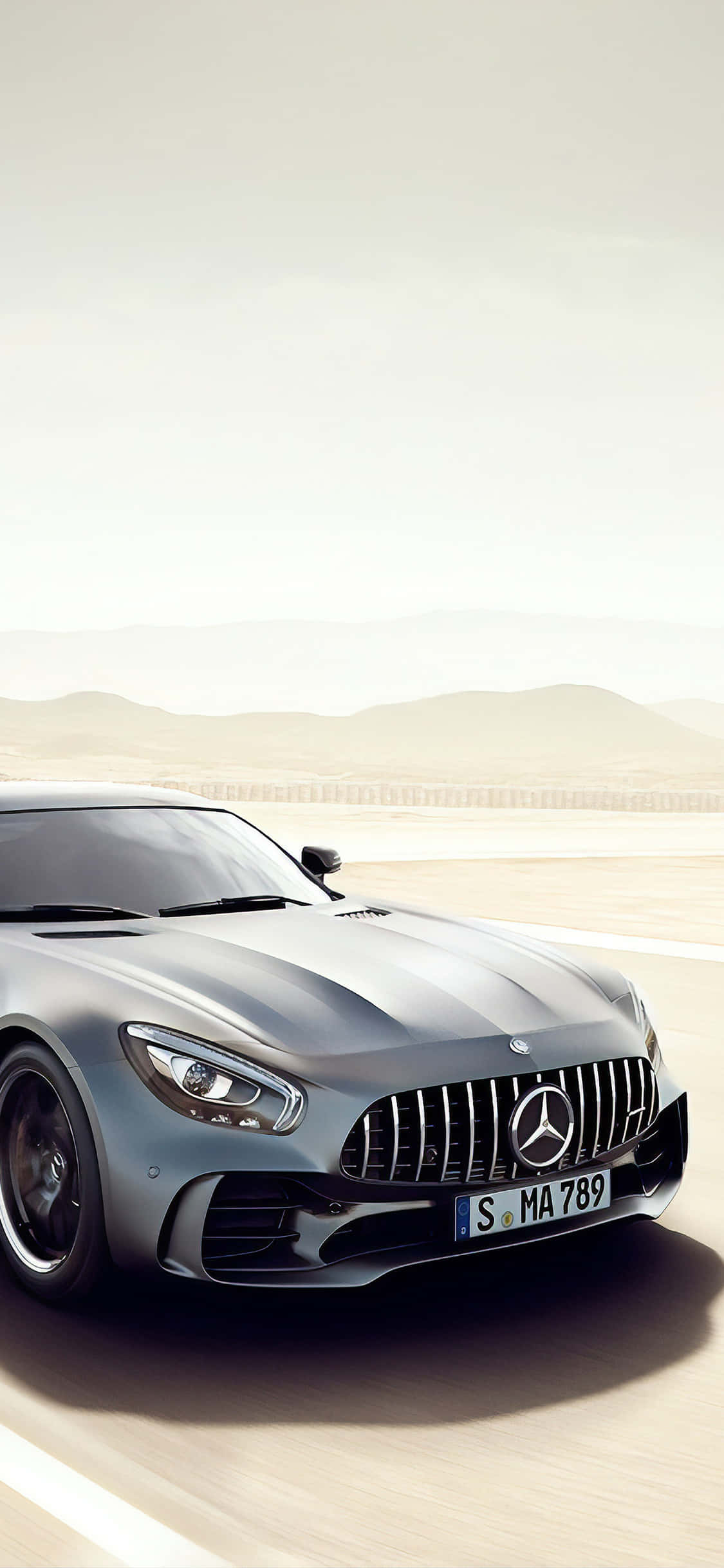 iPhone X Amg Gt-r 桌布 壁紙 — 一輛時尚的銀色 Mercedes-AMG GT 跑車在沙漠公路上奔馳，遠山在朦朧的天空下若隱若現。