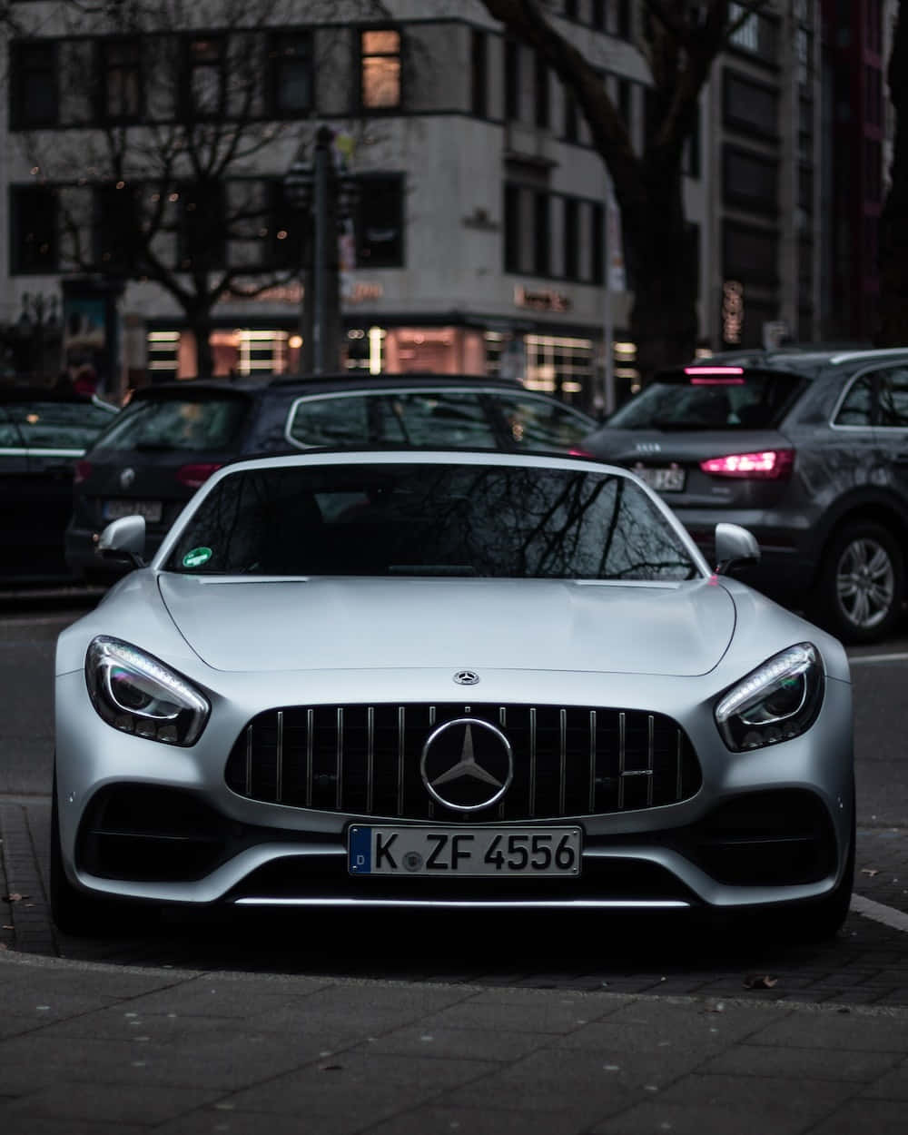 Mercedes 桌布 壁紙 — 一輛時尚的銀色 Mercedes-AMG GT 在黃昏時分停泊在城市街道上，車頭燈閃爍，背景是都市建築。