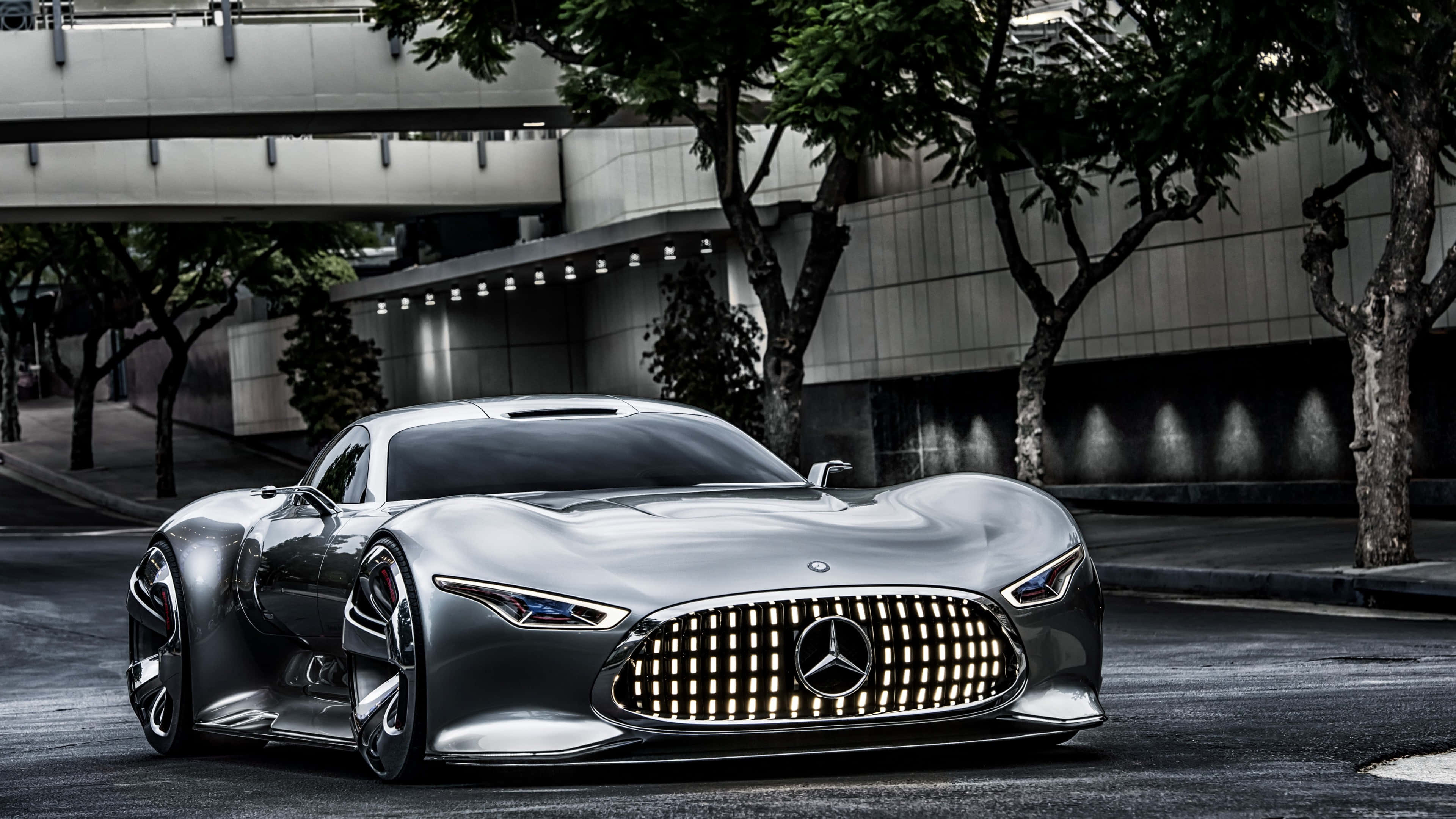 4K 奔馳壁紙 桌布 壁紙 — 一輛時尚的銀色 Mercedes-Benz 概念車在都市街燈下，在枝葉繁茂的樹木和混凝土建築旁閃閃發光。