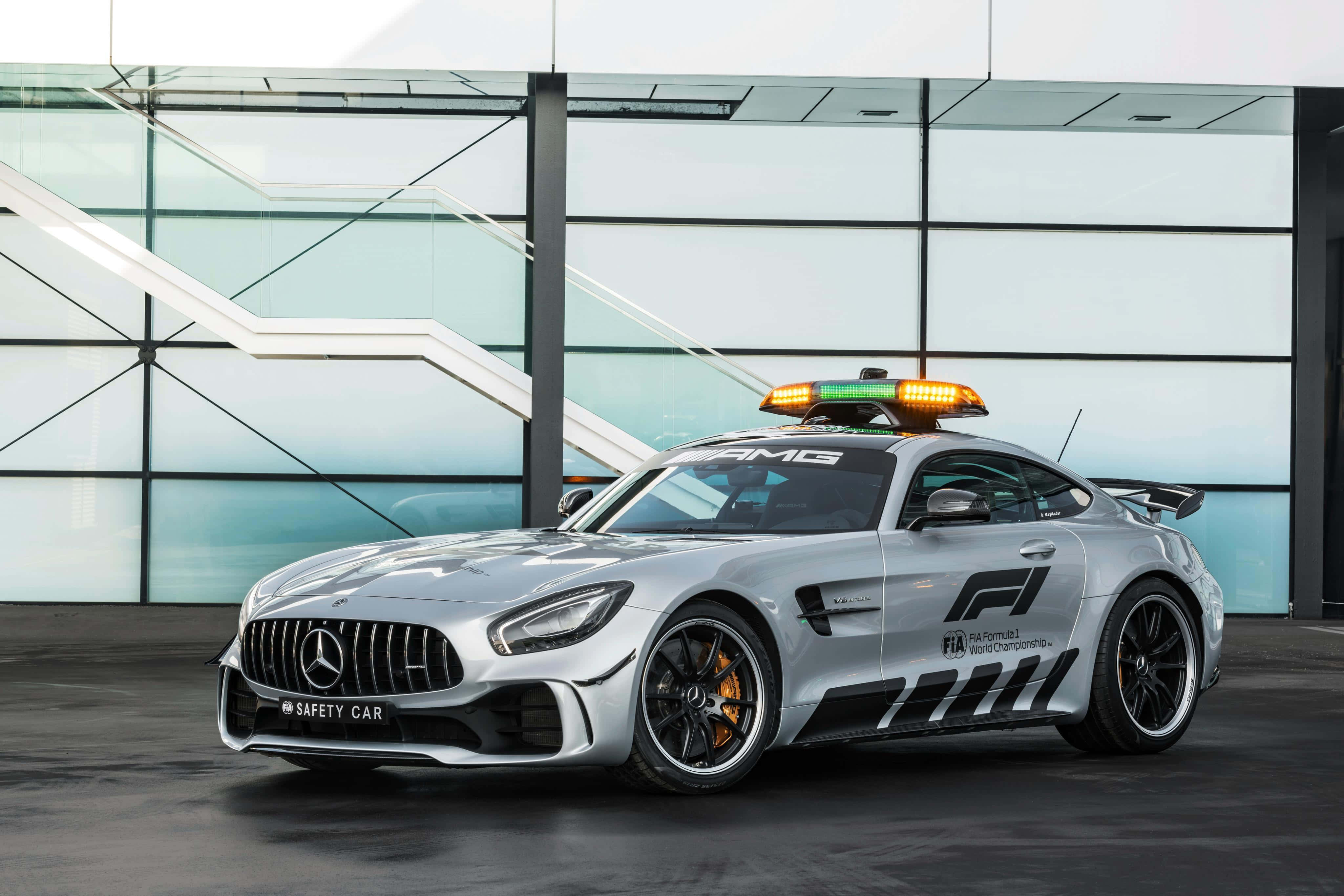 Safety Car 桌布 壁紙 — 一輛時尚的銀色 Mercedes-AMG GT 安全車，閃爍著燈光，停在一棟現代玻璃建築前。