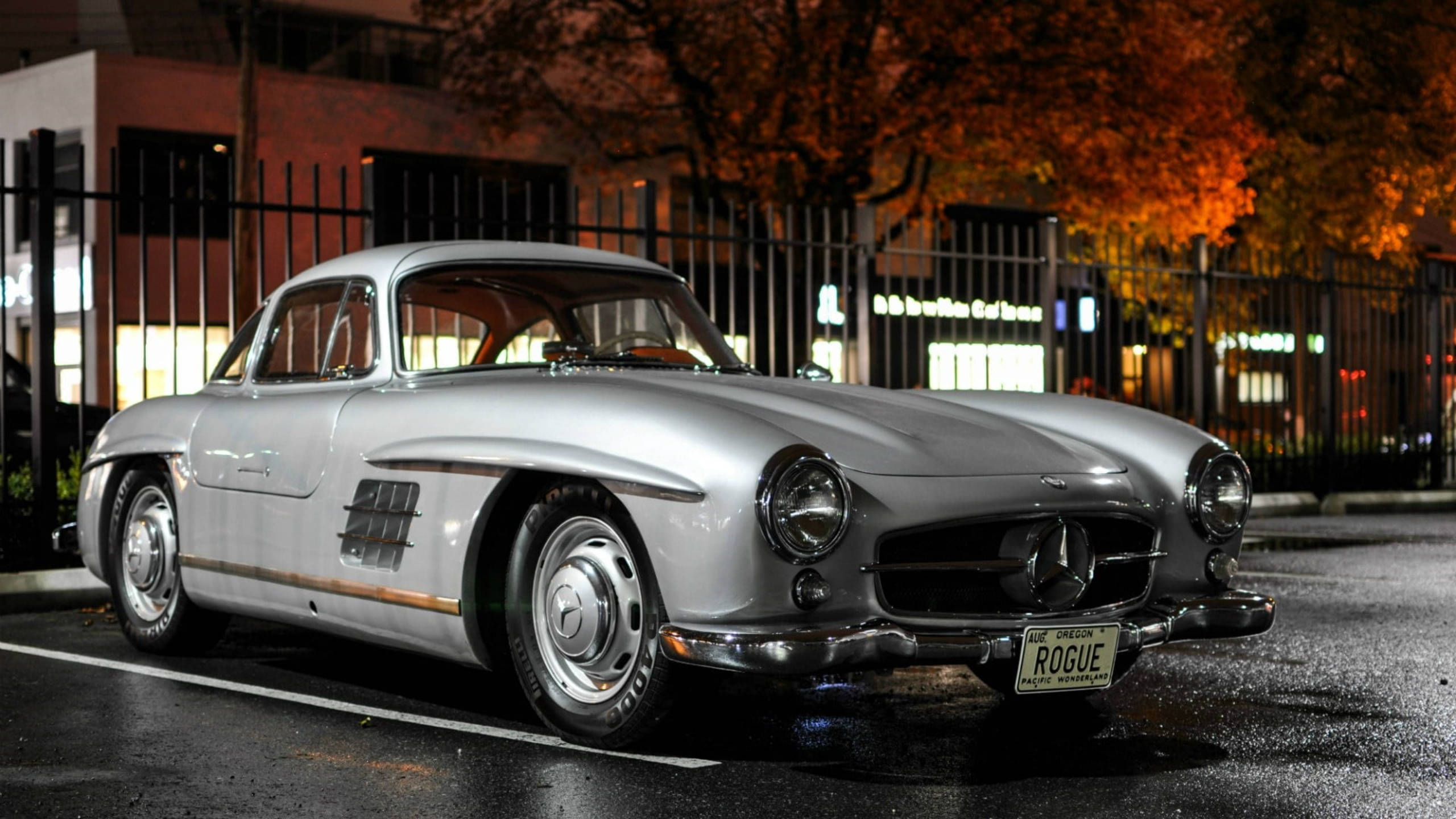 銀色 桌布 壁紙 — 一輛時尚的銀色 vintage Mercedes-Benz 300SL Gullwing 在夜間停泊，秋天的樹木在圍欄後發光。