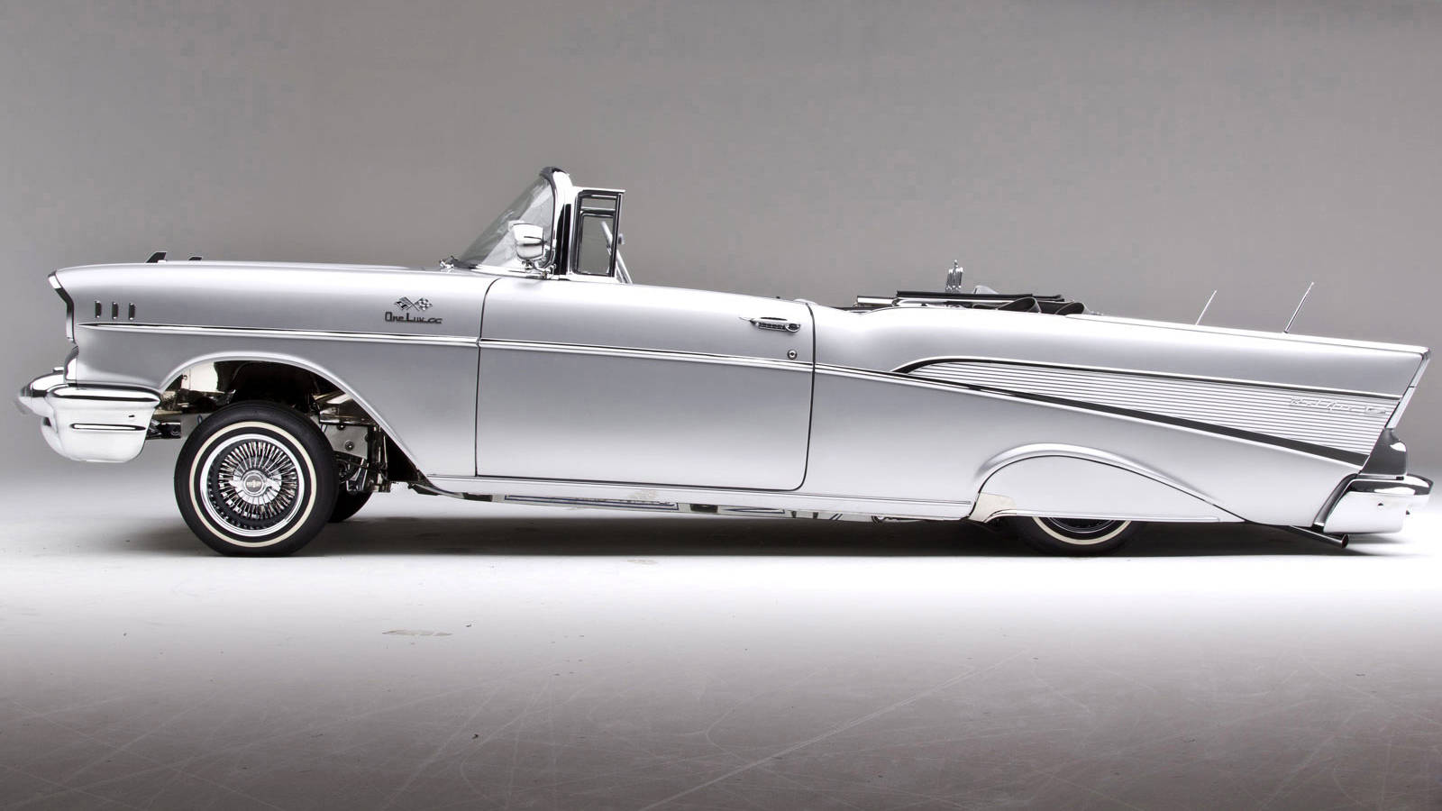 Lowrider 桌布 壁紙 — 一輛時尚的銀色1957年雪佛蘭Bel Air敞篷車，帶有鍍鉻裝飾和白牆輪胎，在攝影棚環境中展示。