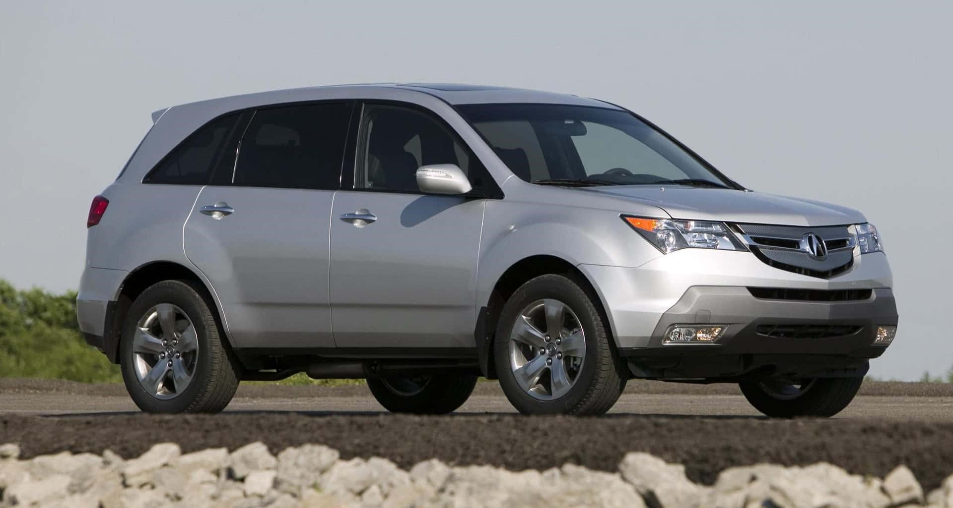 Acura Mdx 桌布 壁紙 — 一輛時尚的銀色Acura SUV停在碎石上，背景有綠植，天空晴朗。
