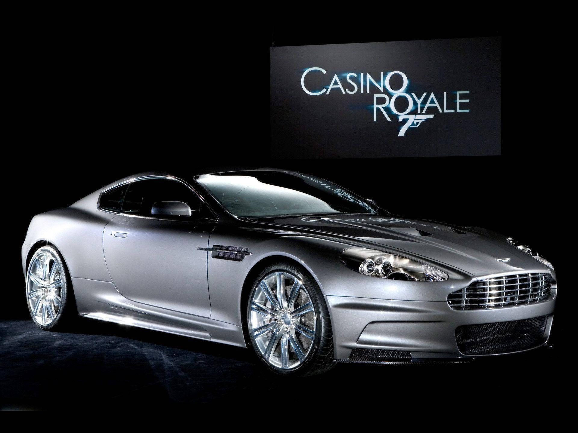 電影動圖 桌布 壁紙 — 一輛時尚的銀色Aston Martin DBS跑車在攝影棚燈光下閃耀，黑色背景襯托著Casino Royale 75標誌螢幕。