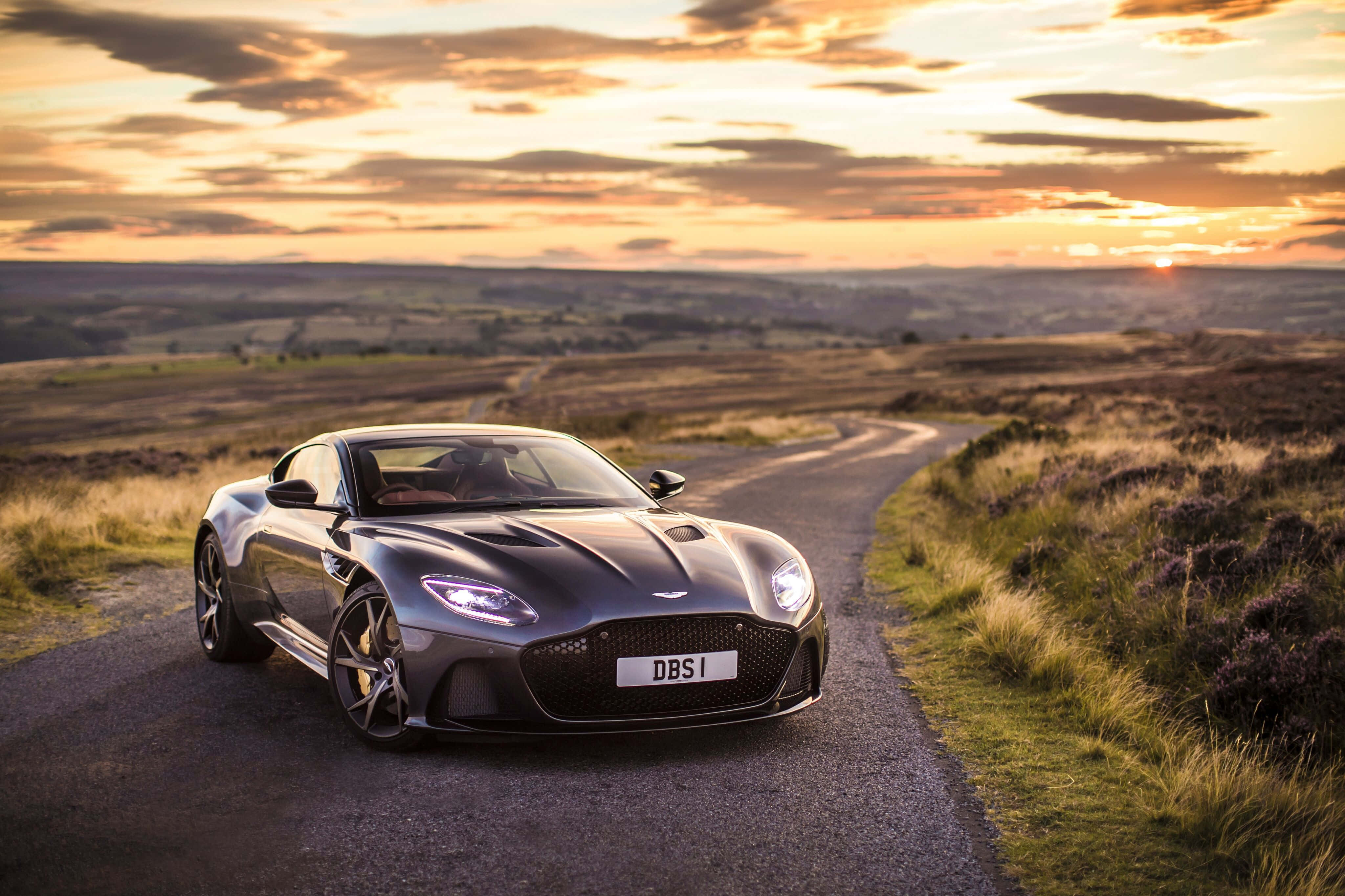 Aston Martin V12 Vantage 桌布 壁紙 — 一輛時尚的銀色Aston Martin DBS1跑車在金色日落時分沿著蜿蜒的鄉村道路滑行。