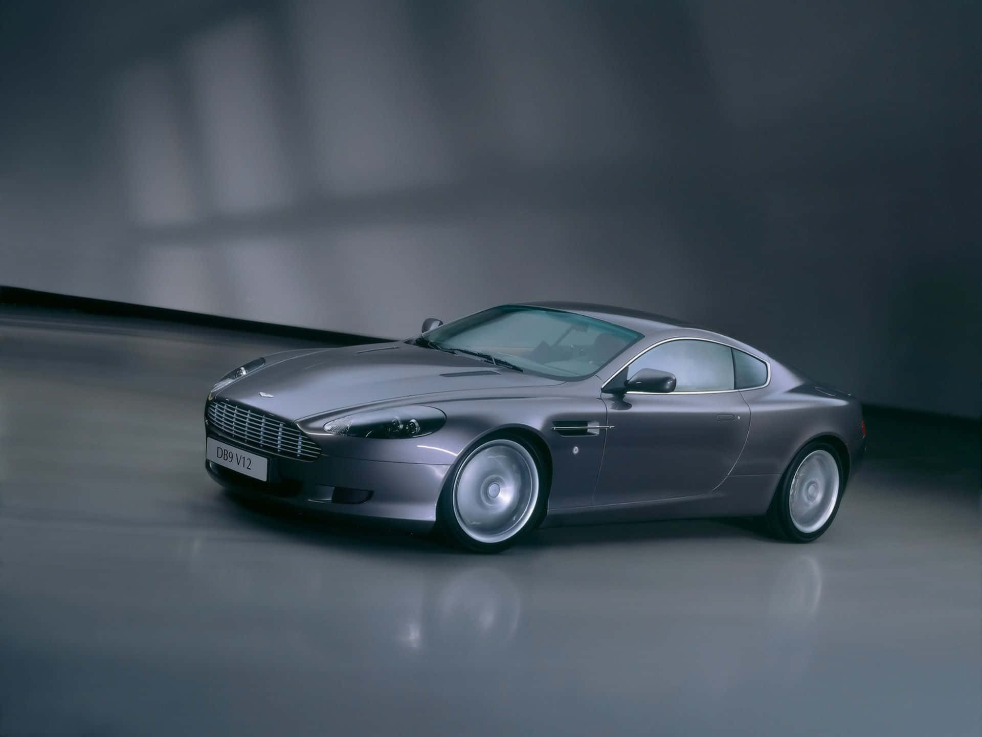 Aston Martin Db9 桌布 壁紙 — 一輛時尚的銀色Aston Martin DB9 V12雙門跑車在攝影棚燈光下，在反光的灰色地板上閃閃發光。