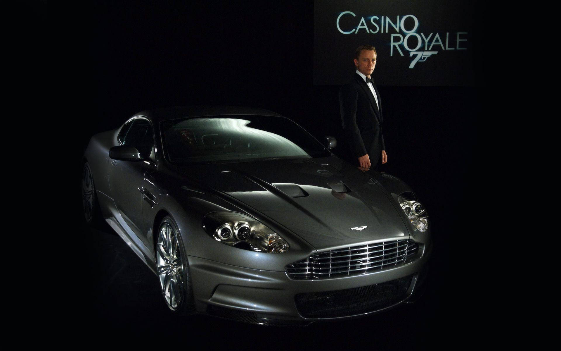 Bond 桌布 壁紙 — 一輛時尚的銀色Aston Martin DBS在聚光燈下閃閃發光，丹尼爾·克雷格站在旁邊，背景是黑色，帶有Casino Royale標誌。