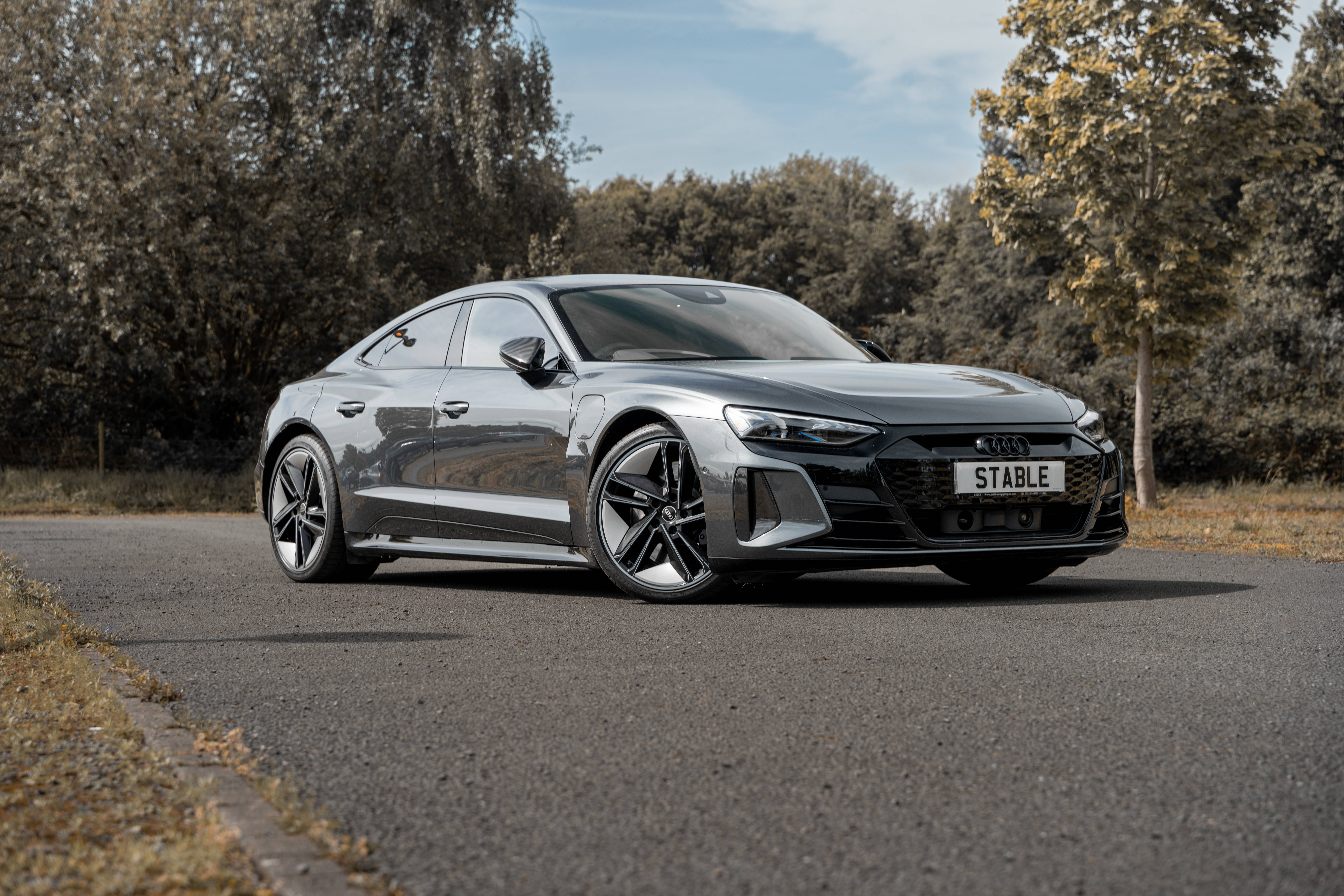 Rs 桌布 壁紙 — 一輛時尚的銀色Audi e-tron GT電動轎跑車，停放在寧靜的柏油路上，兩旁是秋日樹木，天空清澈湛藍。