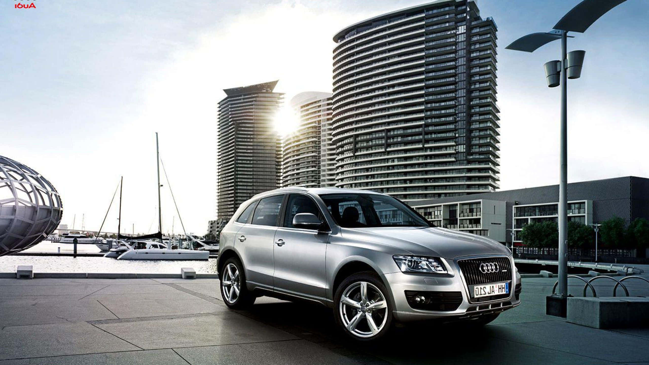 Audi Sq5 桌布 壁紙 — 一輛時尚的銀色Audi Q7 SUV停放在現代海濱廣場上，背景有彎曲的玻璃塔和帆船。