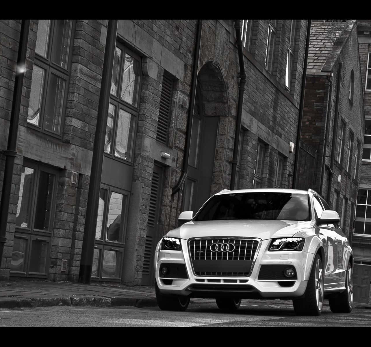 Audi Q5 桌布 壁紙 — 一輛時尚的銀色Audi SUV停放在一條狹窄的鵝卵石街道上，兩旁是歷史悠久的石造建築，在柔和的日光下。
