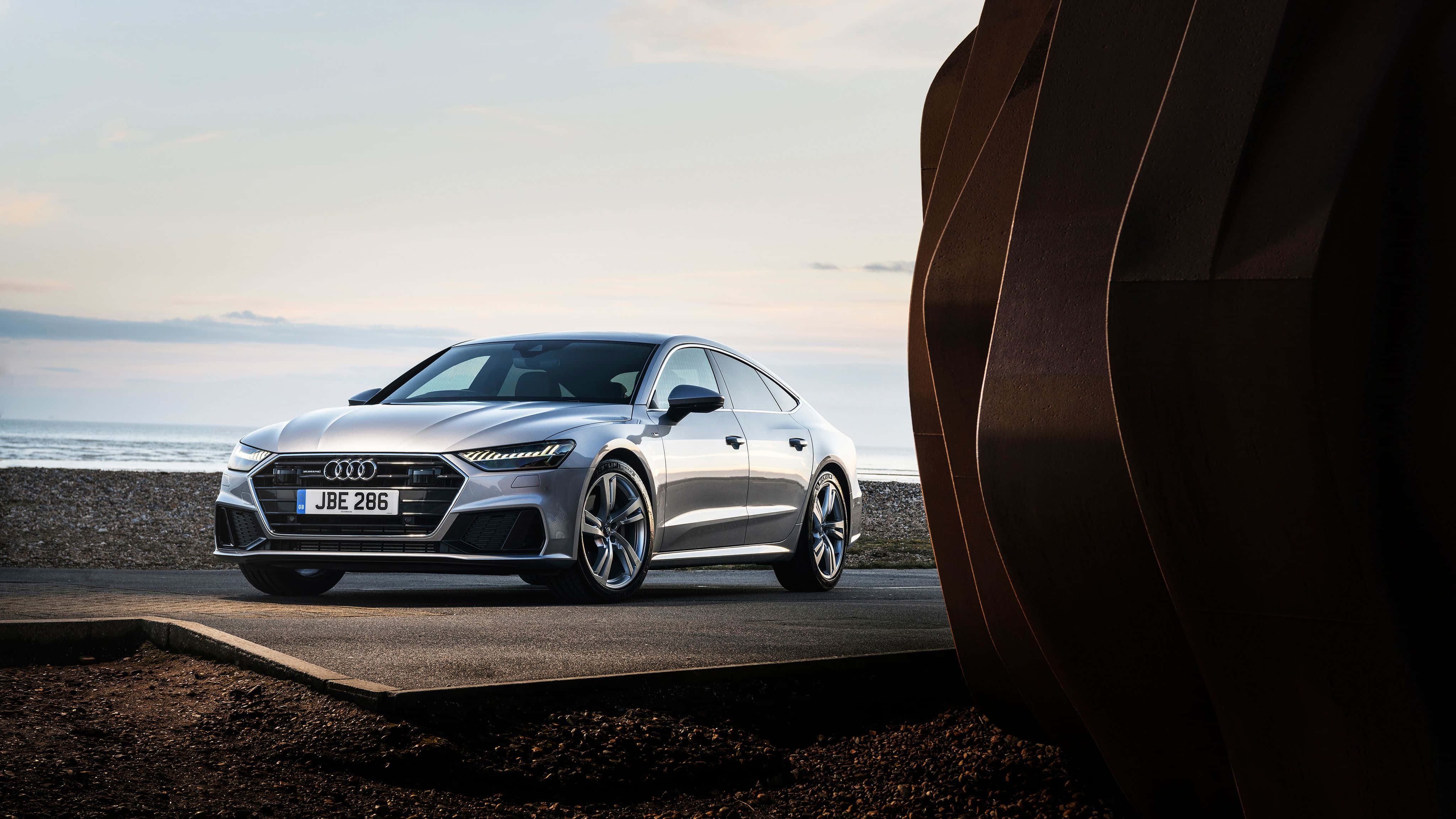 Audi S7 桌布 壁紙 — 一輛時尚的銀色Audi轎車停泊在海岸小徑上，旁邊有一座大型抽象雕塑，背景是海洋和天空。