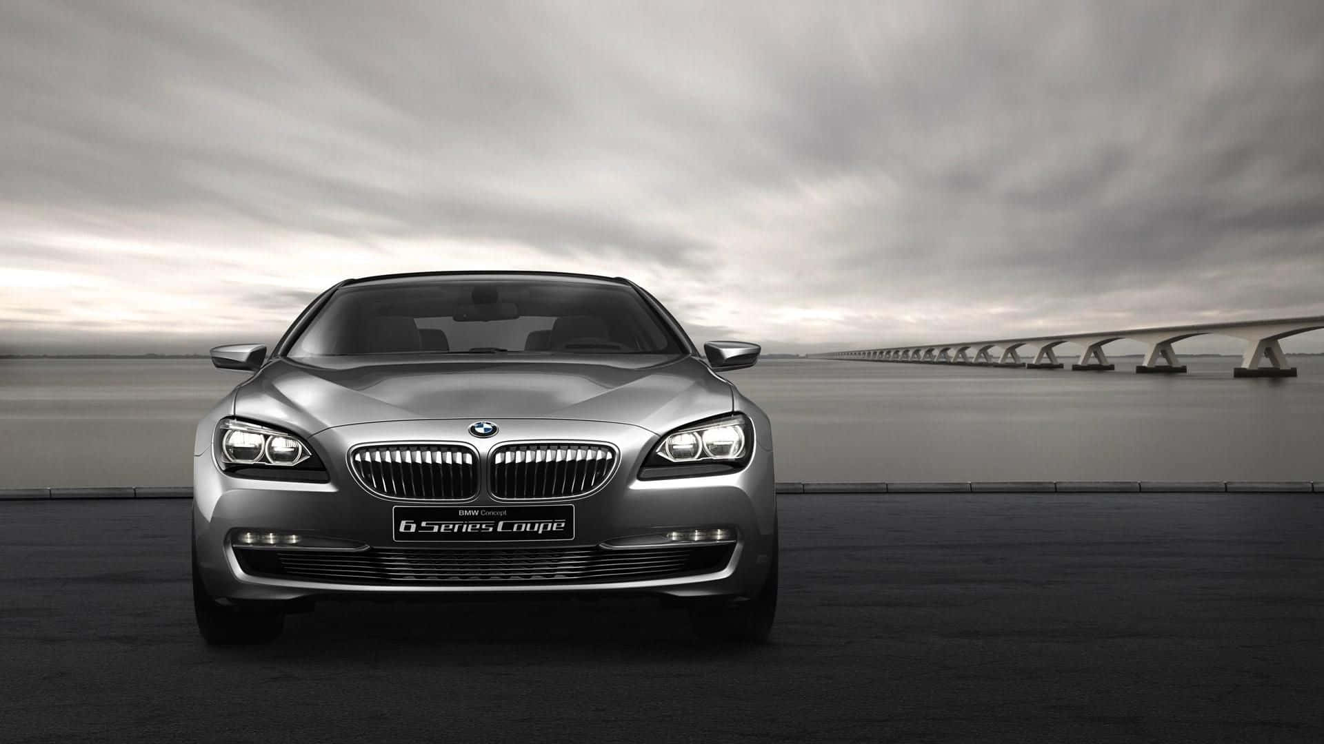 Bmw 6 Series 桌布 壁紙 — 一輛時尚的銀色BMW 6系列轎跑車在黑暗的道路上向前行駛，一條長橋延伸至水面上多雲的天空。