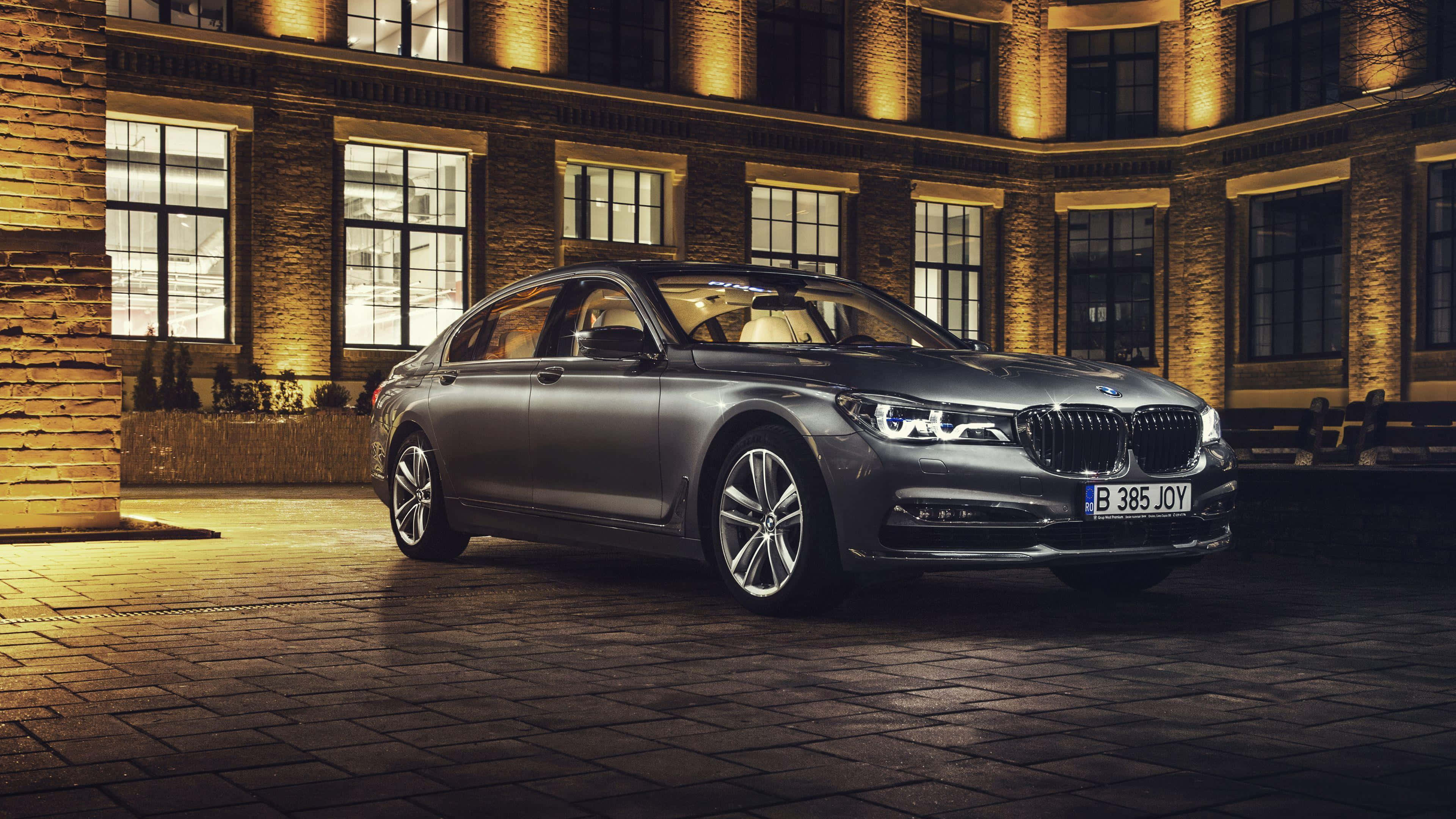 Bmw 7 Series 桌布 壁紙 — 一輛時尚的銀色BMW 7系列轎車在夜間停泊在鵝卵石庭院，被身後歷史悠久的磚造建築散發的溫暖金色燈光照亮。
