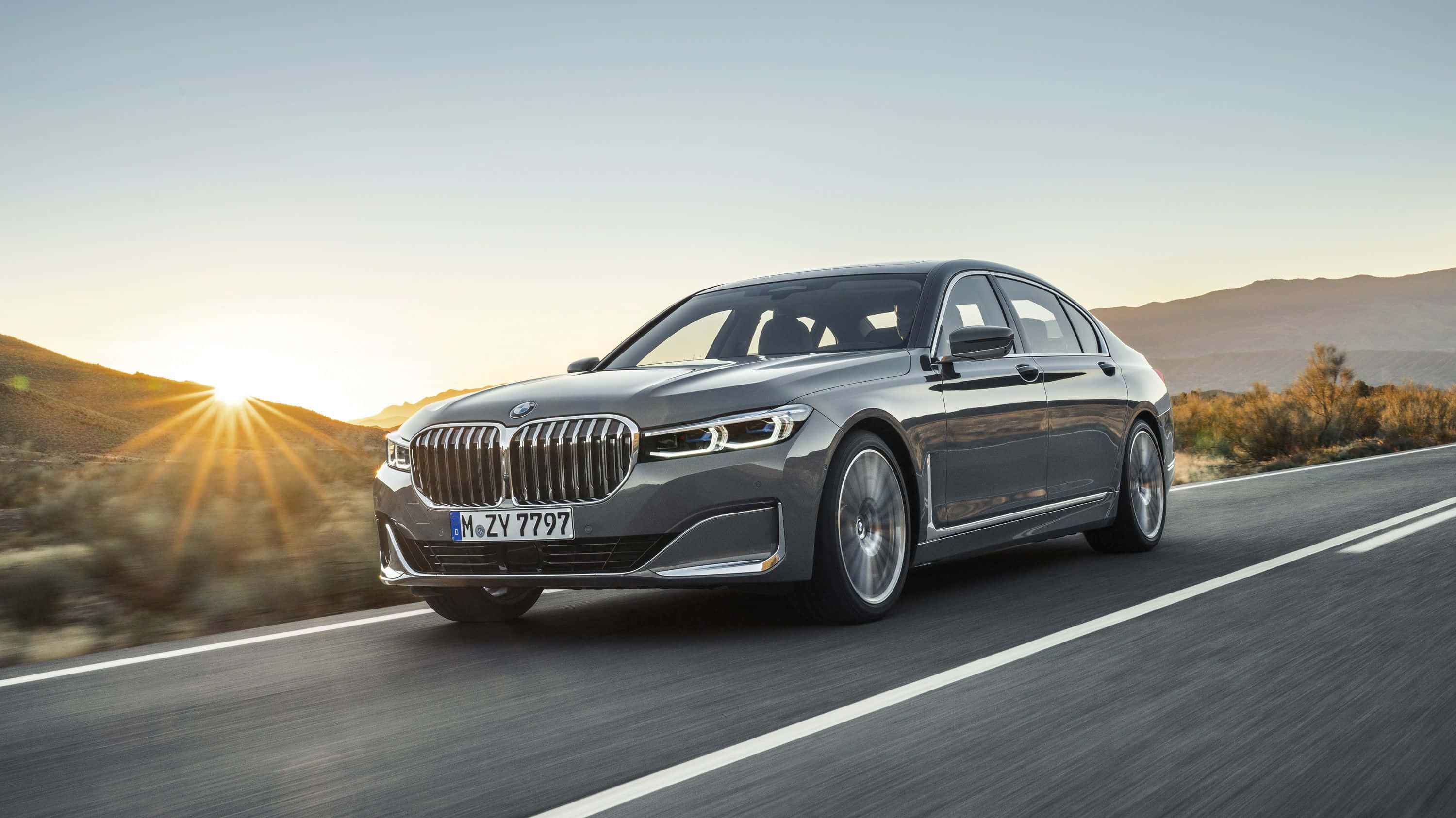 Bmw 7 Series 桌布 壁紙 — 一輛時尚的銀色BMW 7系列轎車在沙漠公路上巡航，夕陽在遠山後落下，灑下金色光芒。