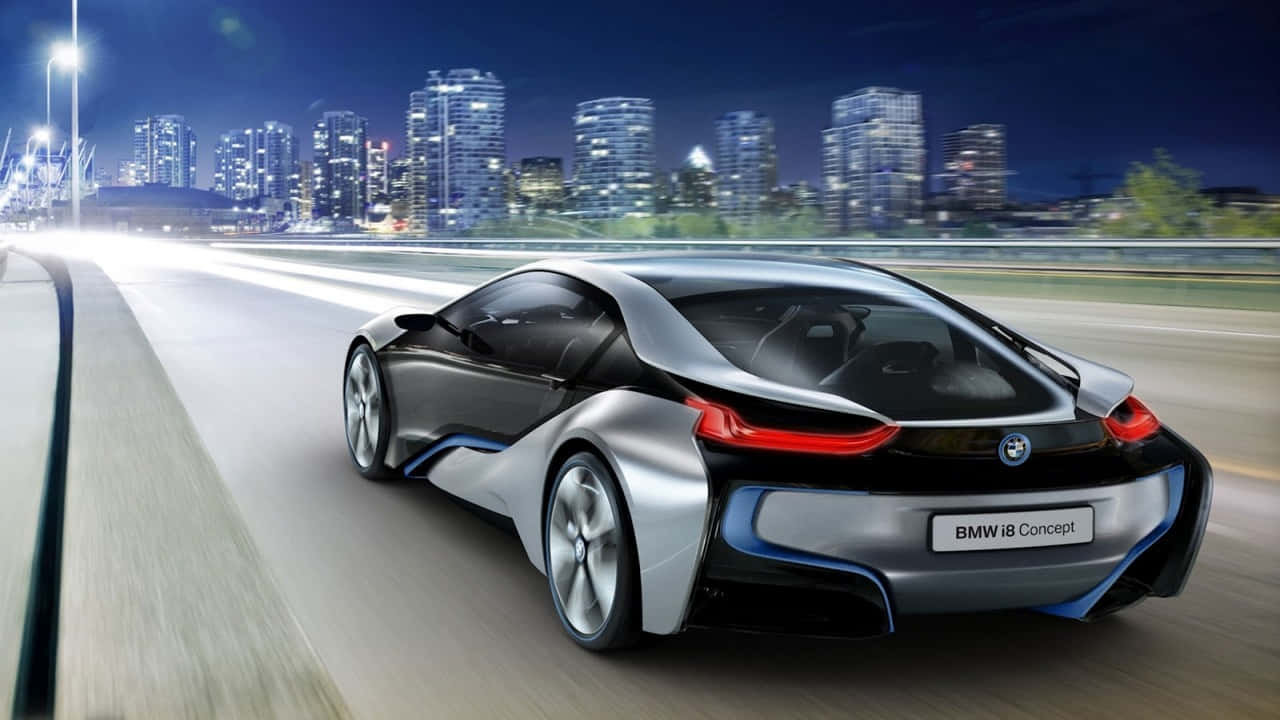 720p Bmw 桌布 壁紙 — 一輛時尚的銀色BMW i8概念車在夜間沿著城市高速公路行駛，背景是閃閃發光的摩天大樓。