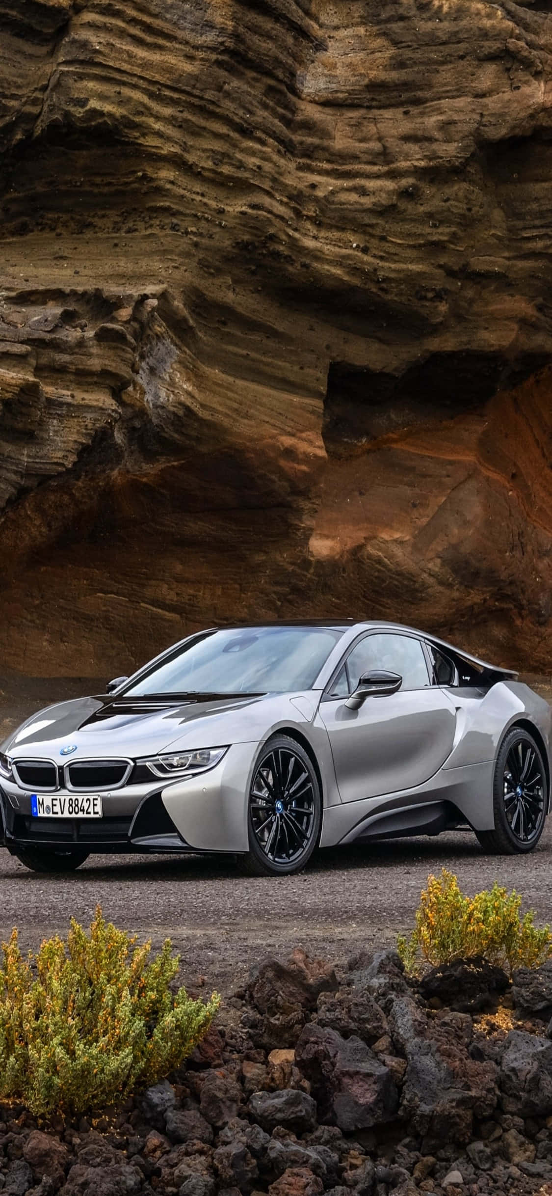 iPhone X Bmw 桌布 壁紙 — 一輛時尚的銀色BMW i8跑車停放在戲劇性的層狀岩層下，周圍有稀疏的綠色灌木。